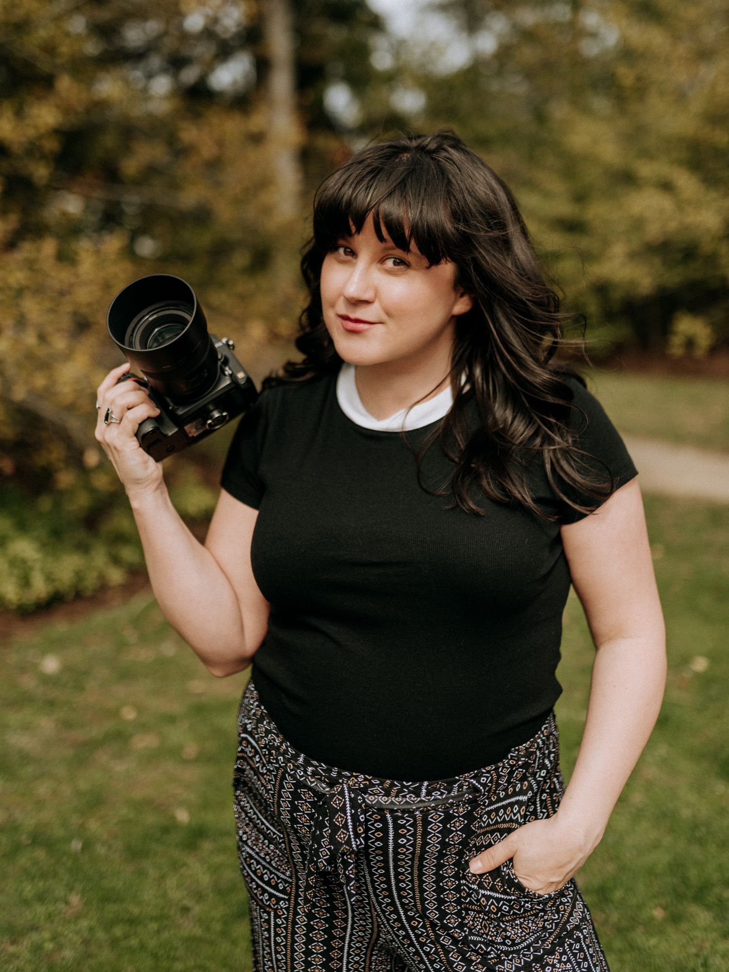 Trista Renee Photography, Female photographer holding a camera, wearing black top and patterned pants, standing in a park.