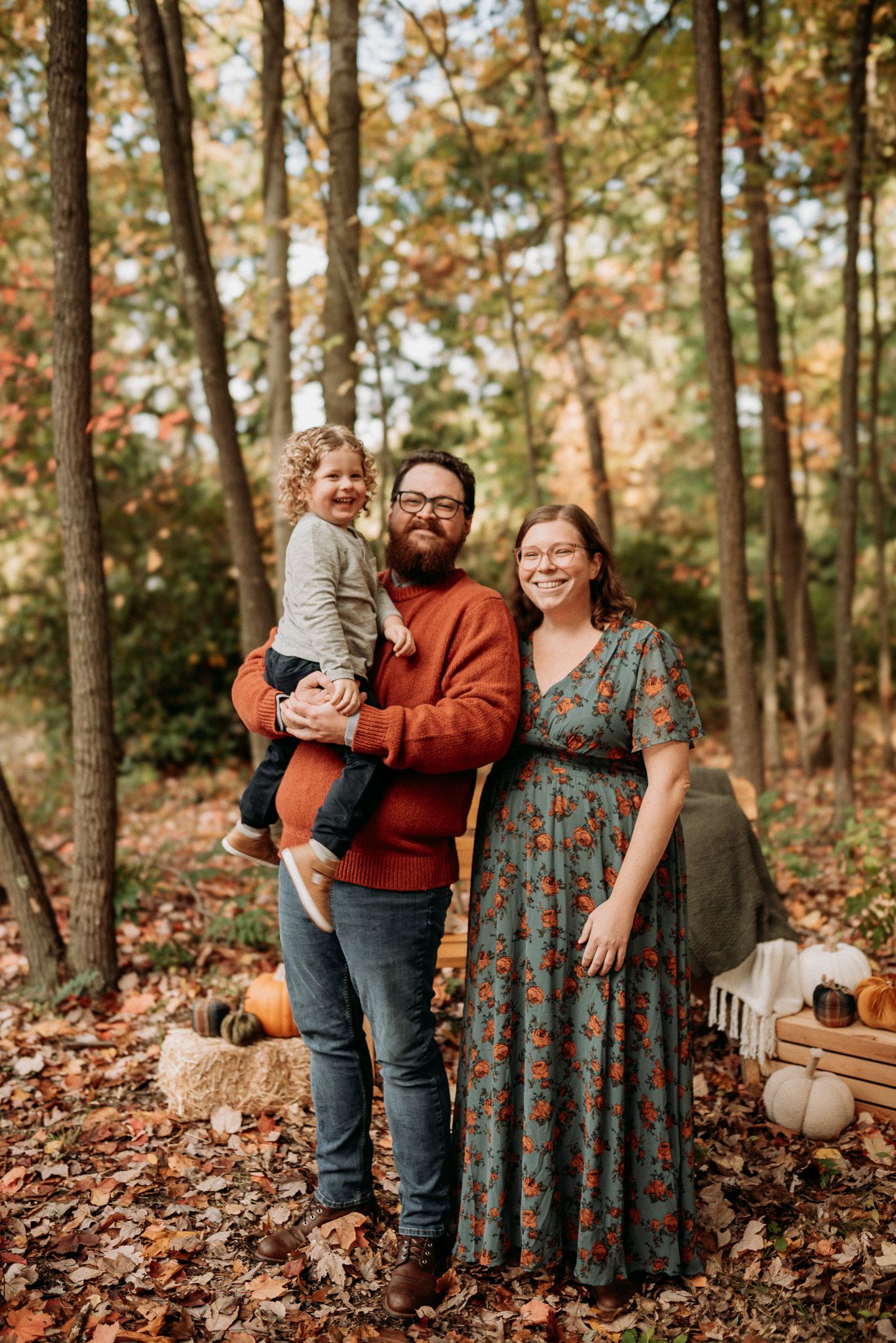 Family of three posing in autumn forest with fall foliage, pumpkins, and seasonal decor during outdoor photo session.