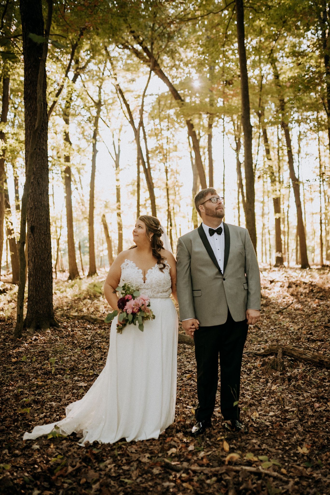 Bride and groom holding hands in sunlit forest, she in lace gown with bouquet, he in gray tuxedo.