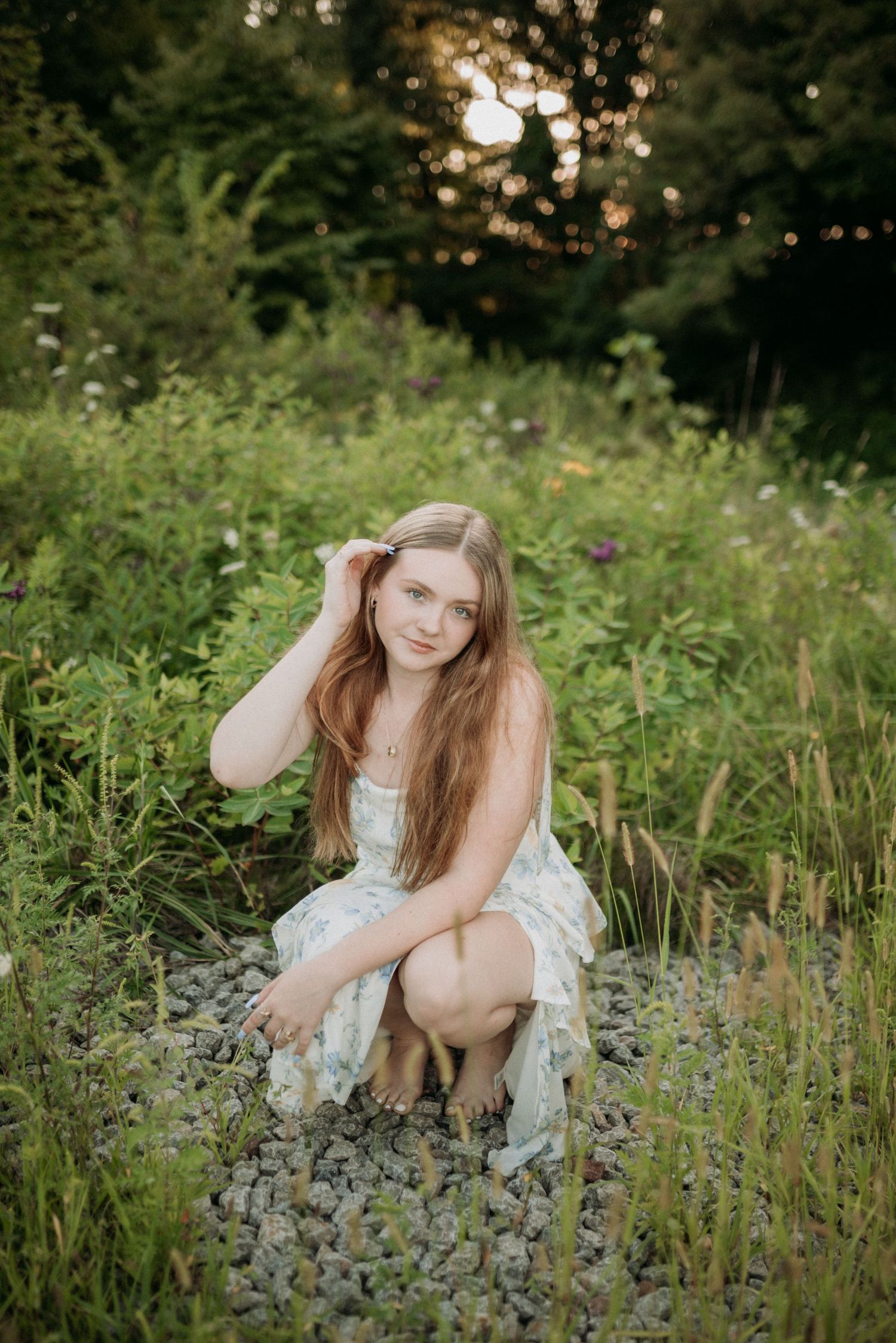Young woman in floral dress crouching on gravel path surrounded by lush green wildflowers at golden hour.