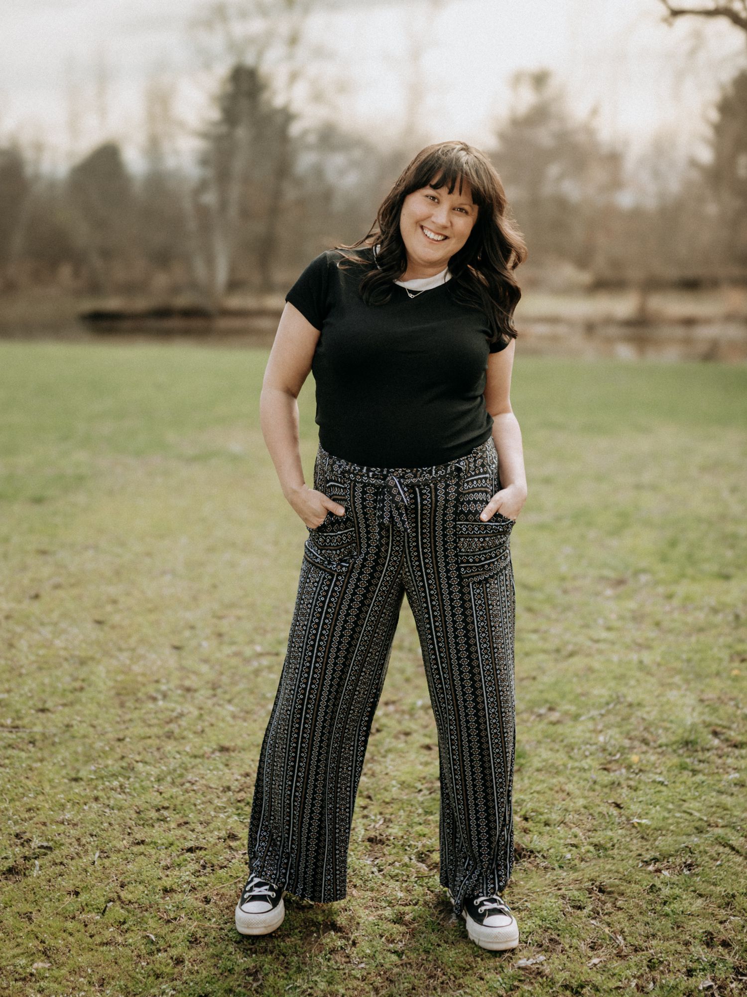 Trista Renee Photography smiling in black top and patterned wide-leg pants, standing outdoors in a grassy park setting.