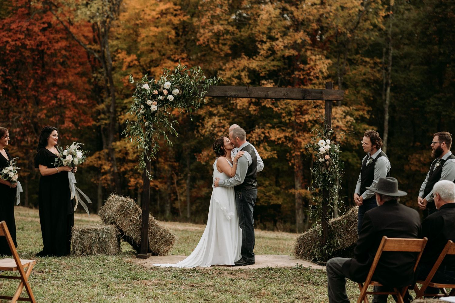 Bride and groom sharing first kiss at rustic outdoor fall wedding ceremony with colorful foliage backdrop.