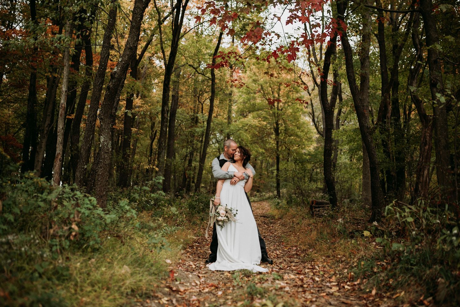 Romantic bride and groom embrace on a leaf-covered forest path surrounded by colorful autumn foliage during fall wedding.