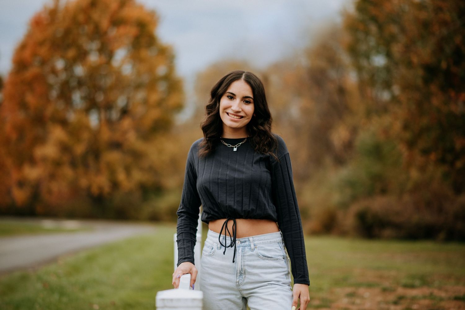 Smiling young brunette woman in black crop top and jeans posing in colorful autumn park setting. Senior Portrait Photography.