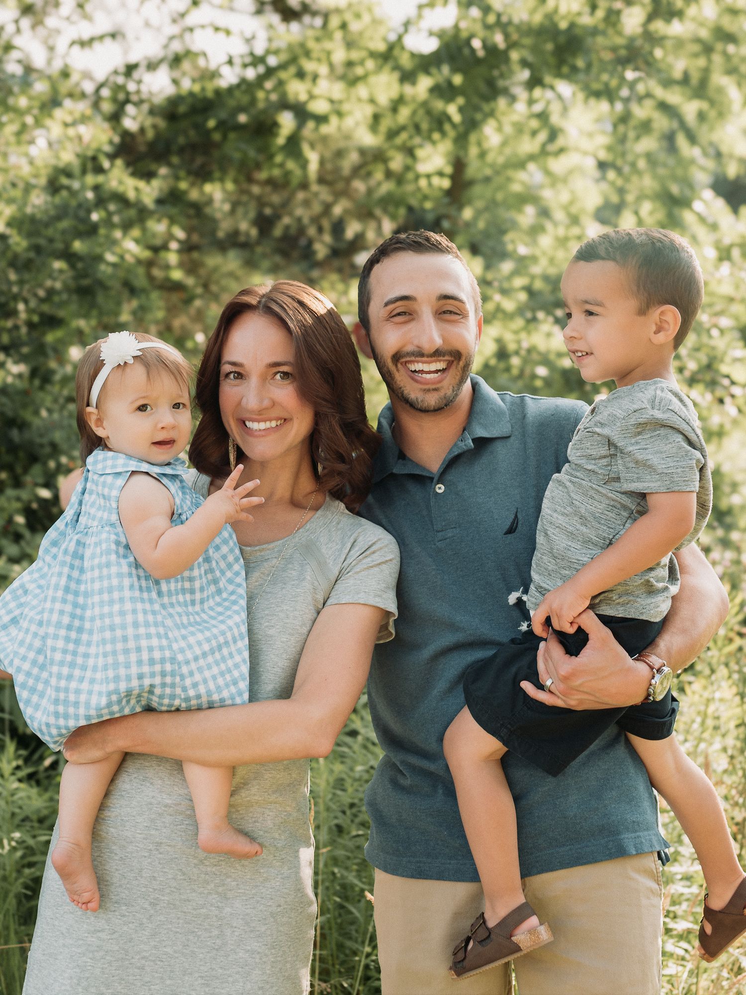 Happy family of four smiles outdoors, parents holding a baby girl in blue dress and toddler boy among green foliage.