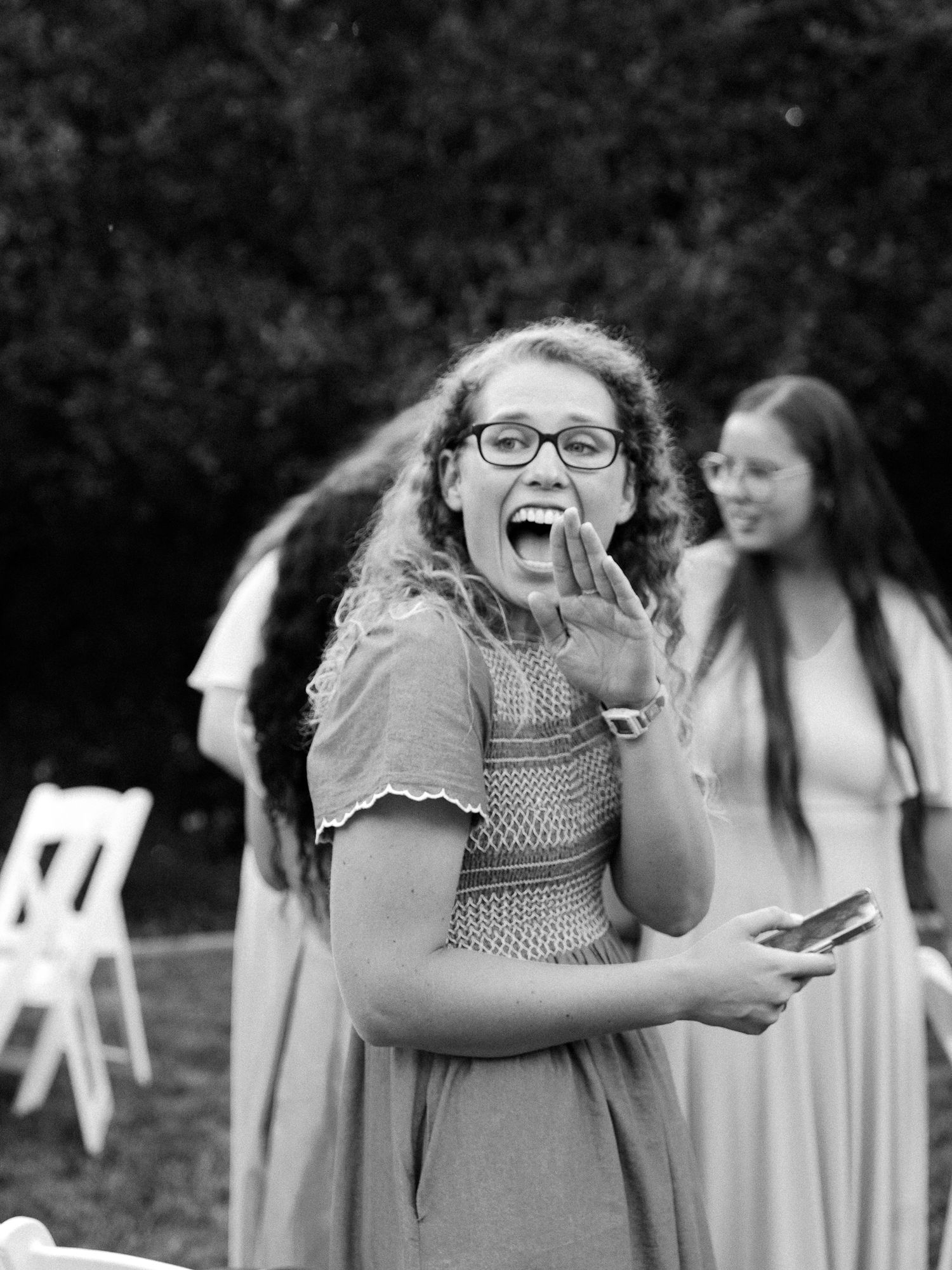 A young woman with curly hair and glasses laughs joyfully outdoors at a garden event in black and white.