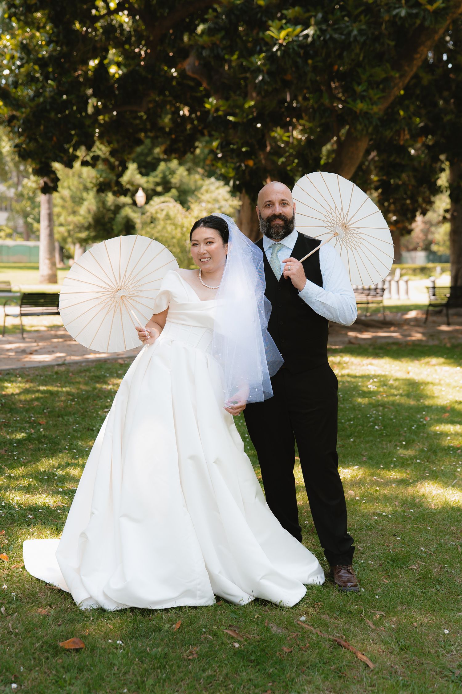 Bride and groom posing with white paper parasols on green grass in a sunlit park on their wedding day.