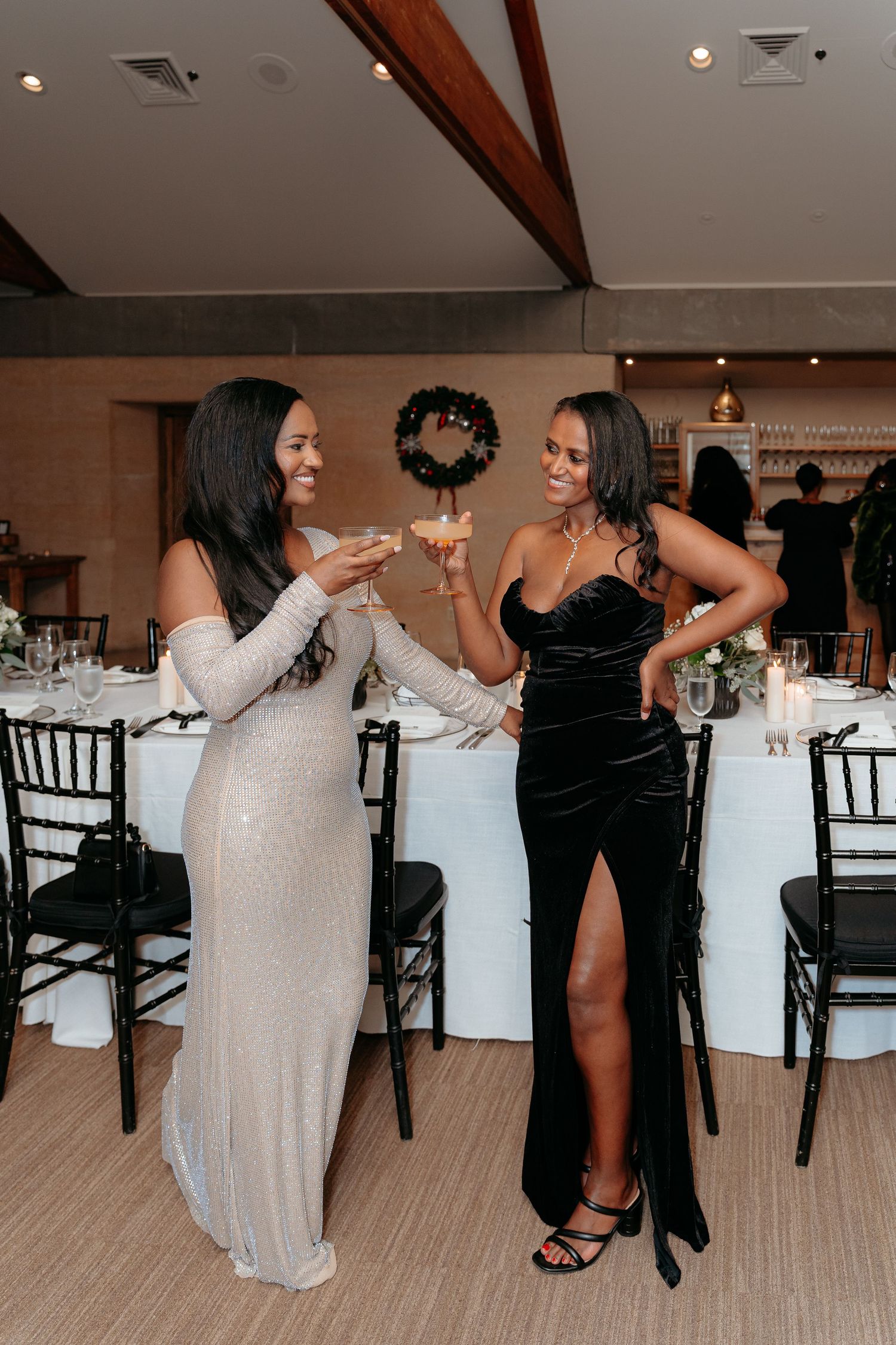 Two women in elegant gowns toasting cocktails at a wedding reception venue.