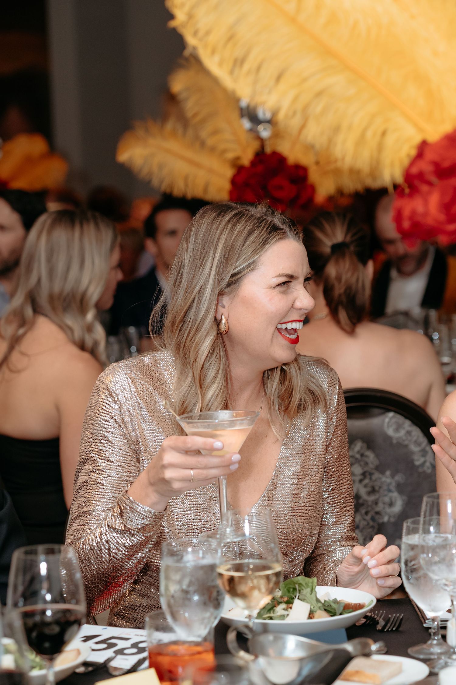 Guest in a sparkly dress laughs while dining at an elegant event with decorative feather centerpieces.