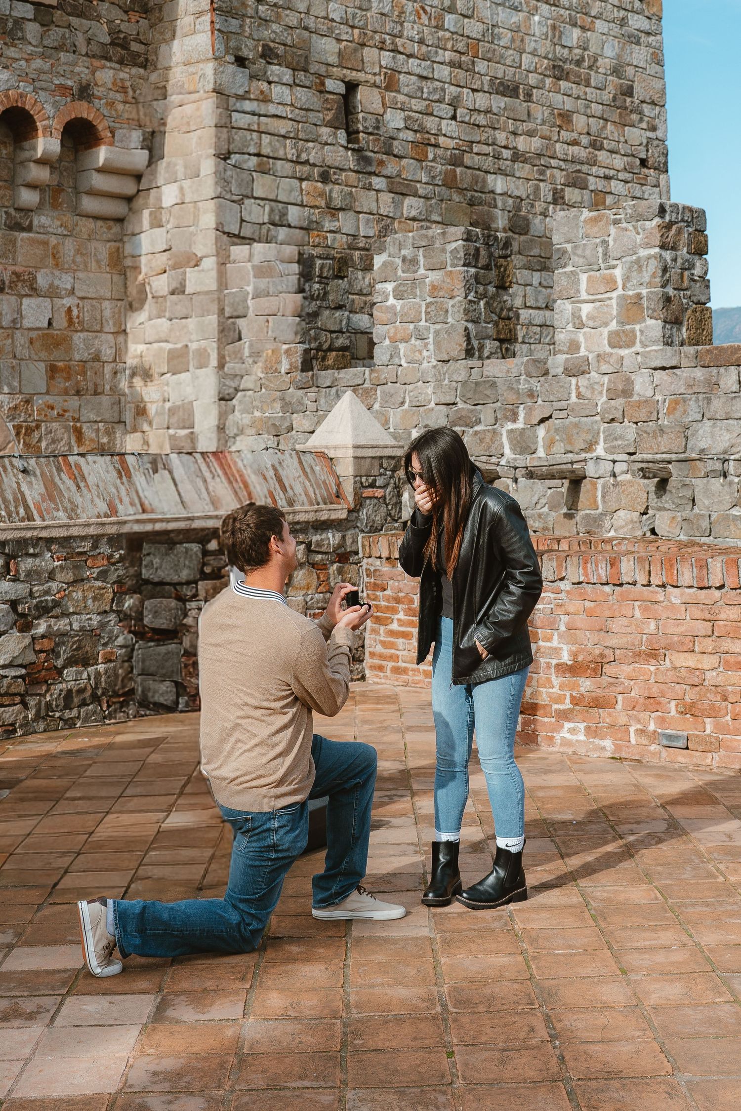 Man proposes on one knee to woman near historic stone castle wall, holding ring box as she covers her mouth in surprise.