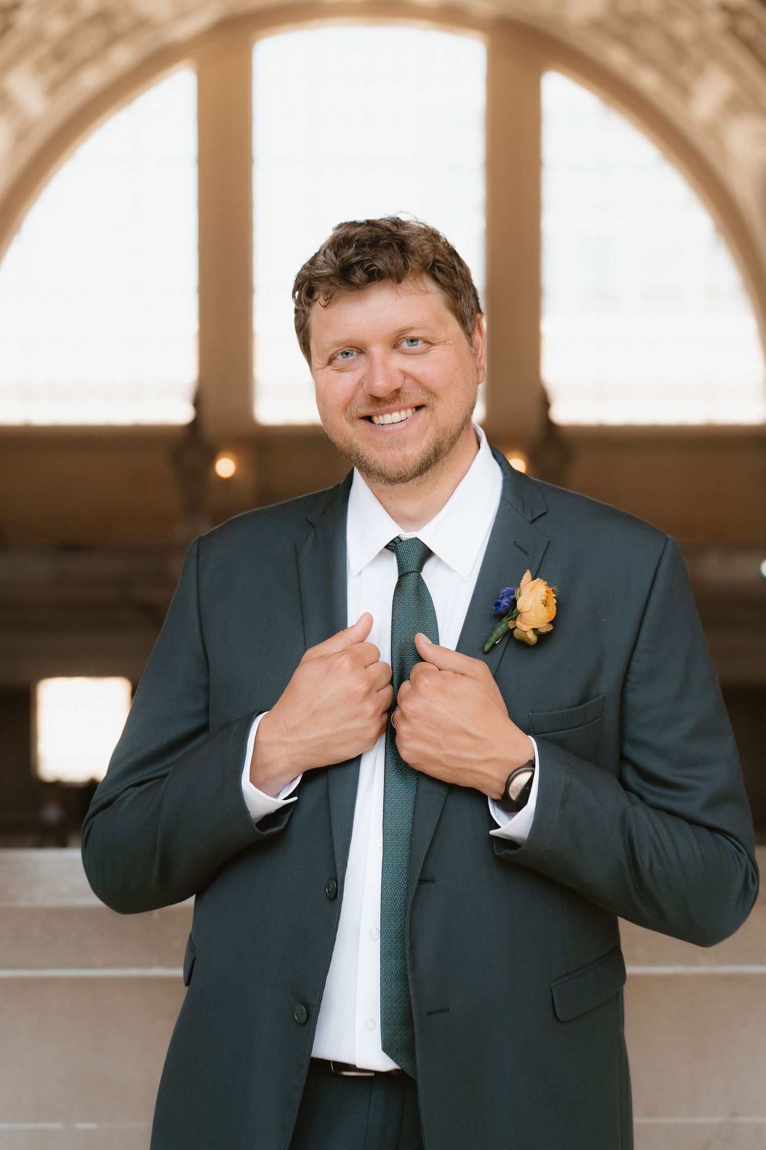 Smiling groom in dark green suit adjusts lapels, wearing green tie and yellow rose boutonniere indoors.
