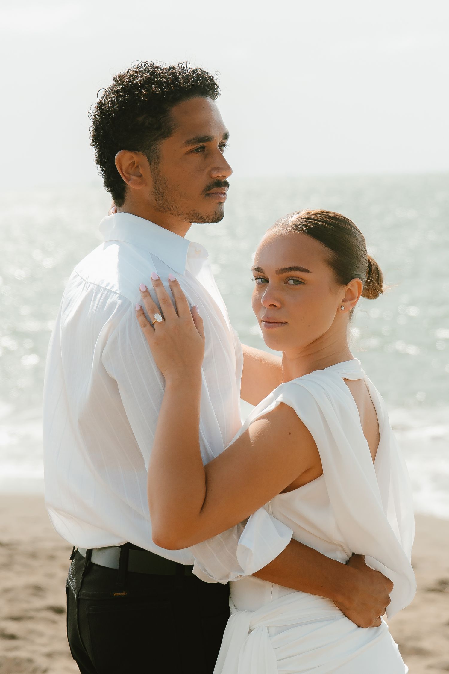 Couple in white attire embracing on a beach, woman wearing engagement ring, ocean backdrop.