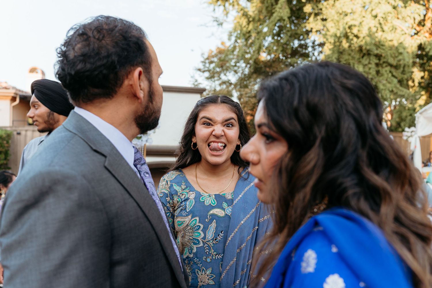 Three people in festive attire chat outdoors, with a woman in blue floral dress making a funny face.