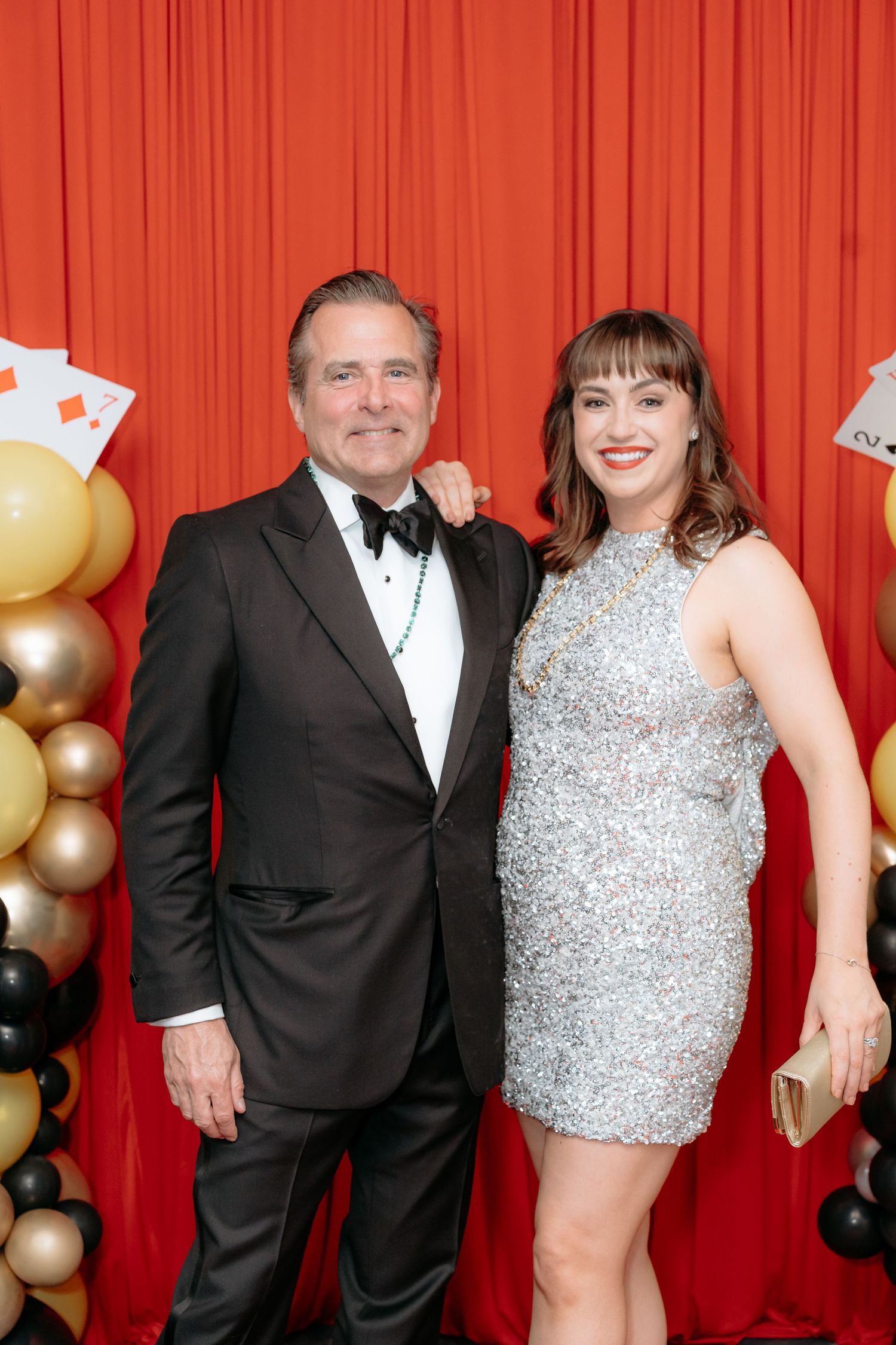 Couple posing at a casino-themed gala, man in tuxedo and woman in silver sequin dress before red curtain backdrop.