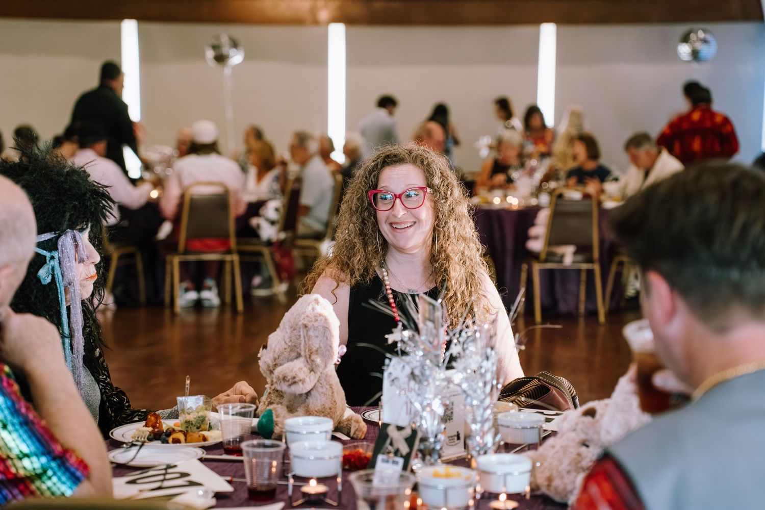 Woman with curly hair and red glasses smiling at a decorated banquet table during a festive costume party event.