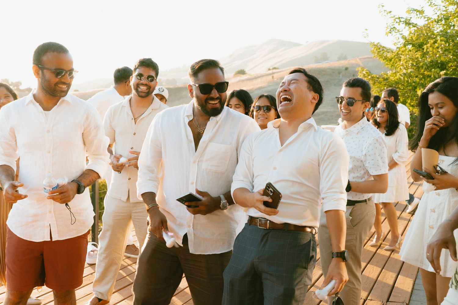 Group of men in white shirts laughing together at an outdoor sunset celebration event.