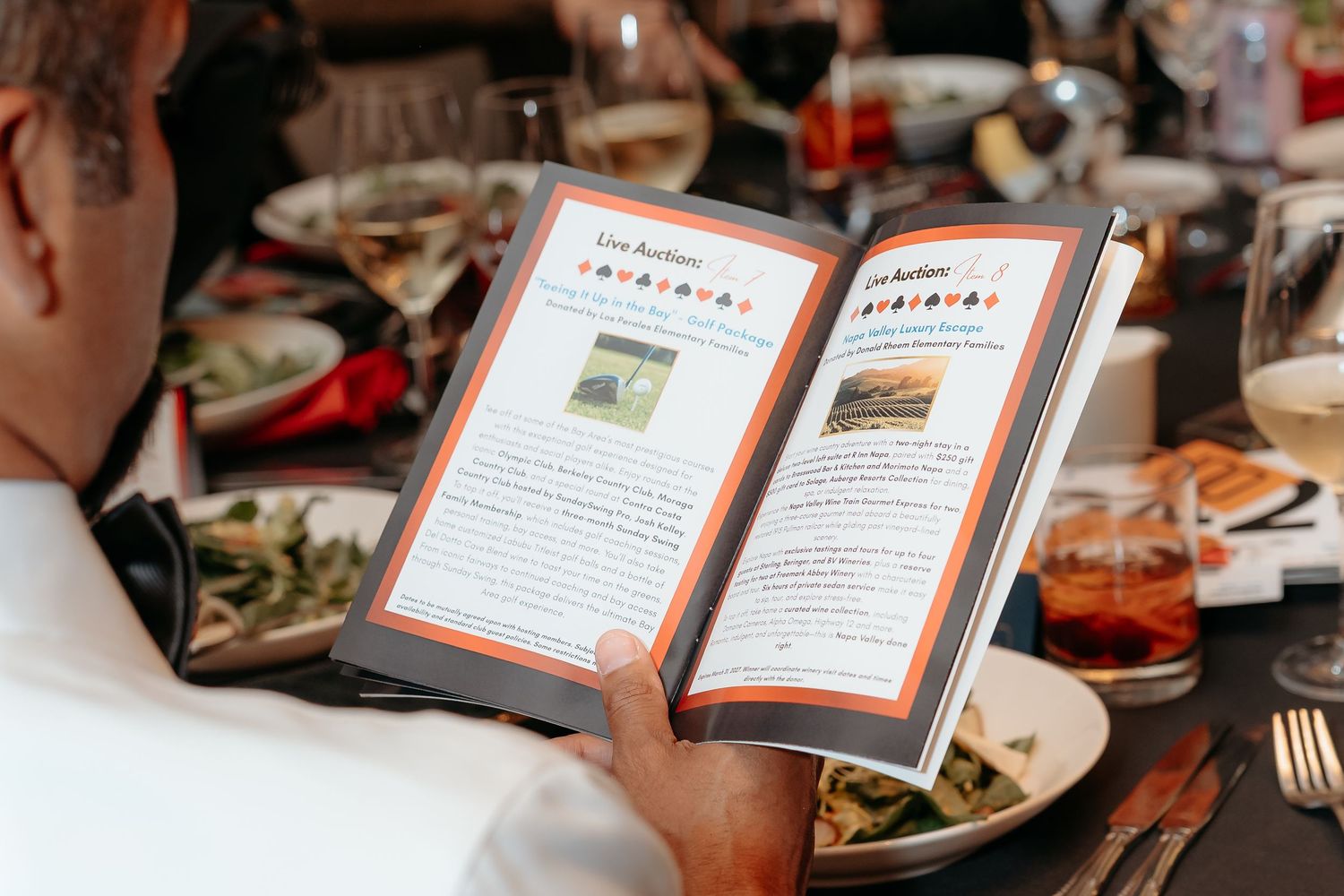 Man reading an open menu book at a restaurant dining table with plates and glasses in the background.