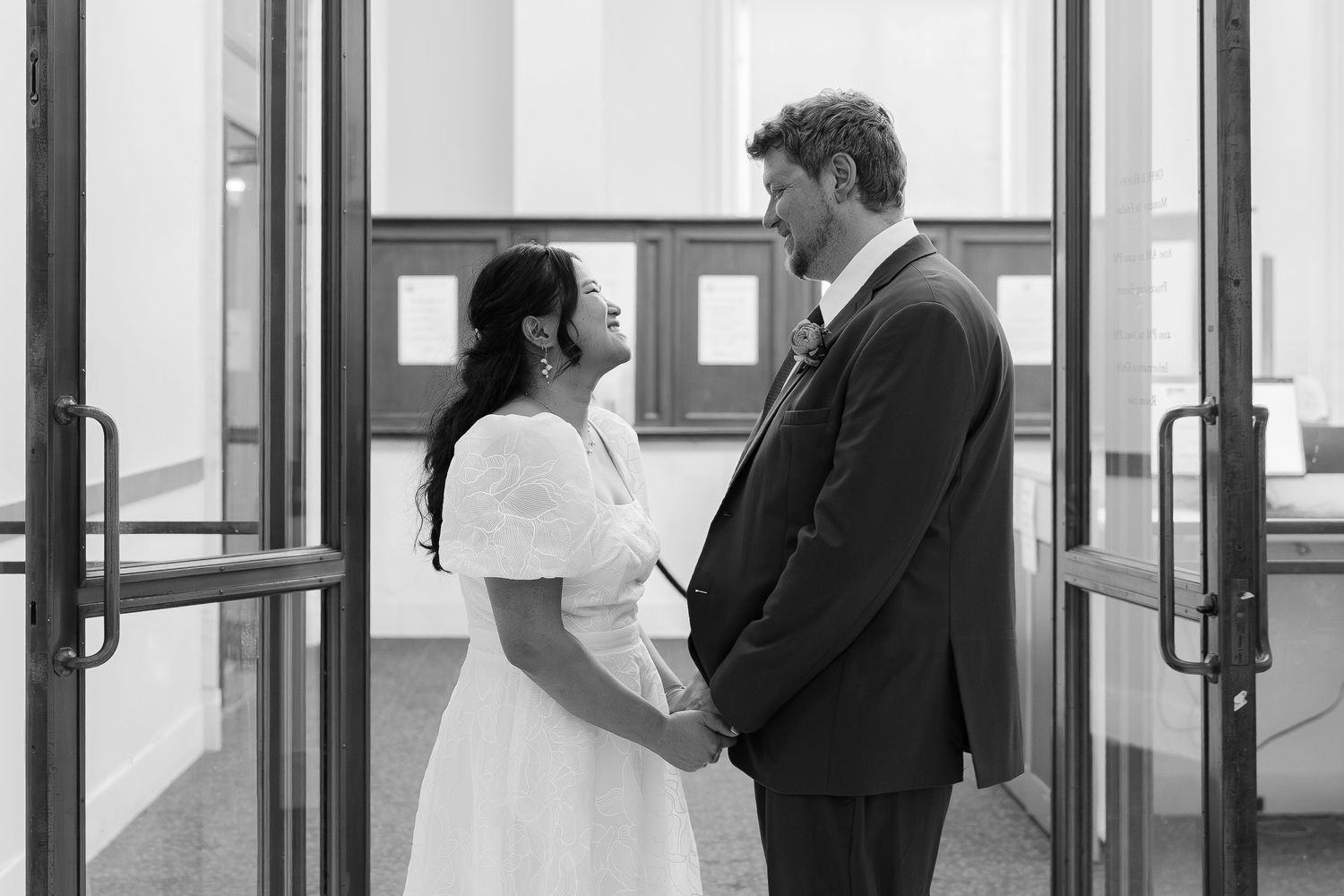 Black and white photo of a couple in wedding attire holding hands and facing each other in a doorway.