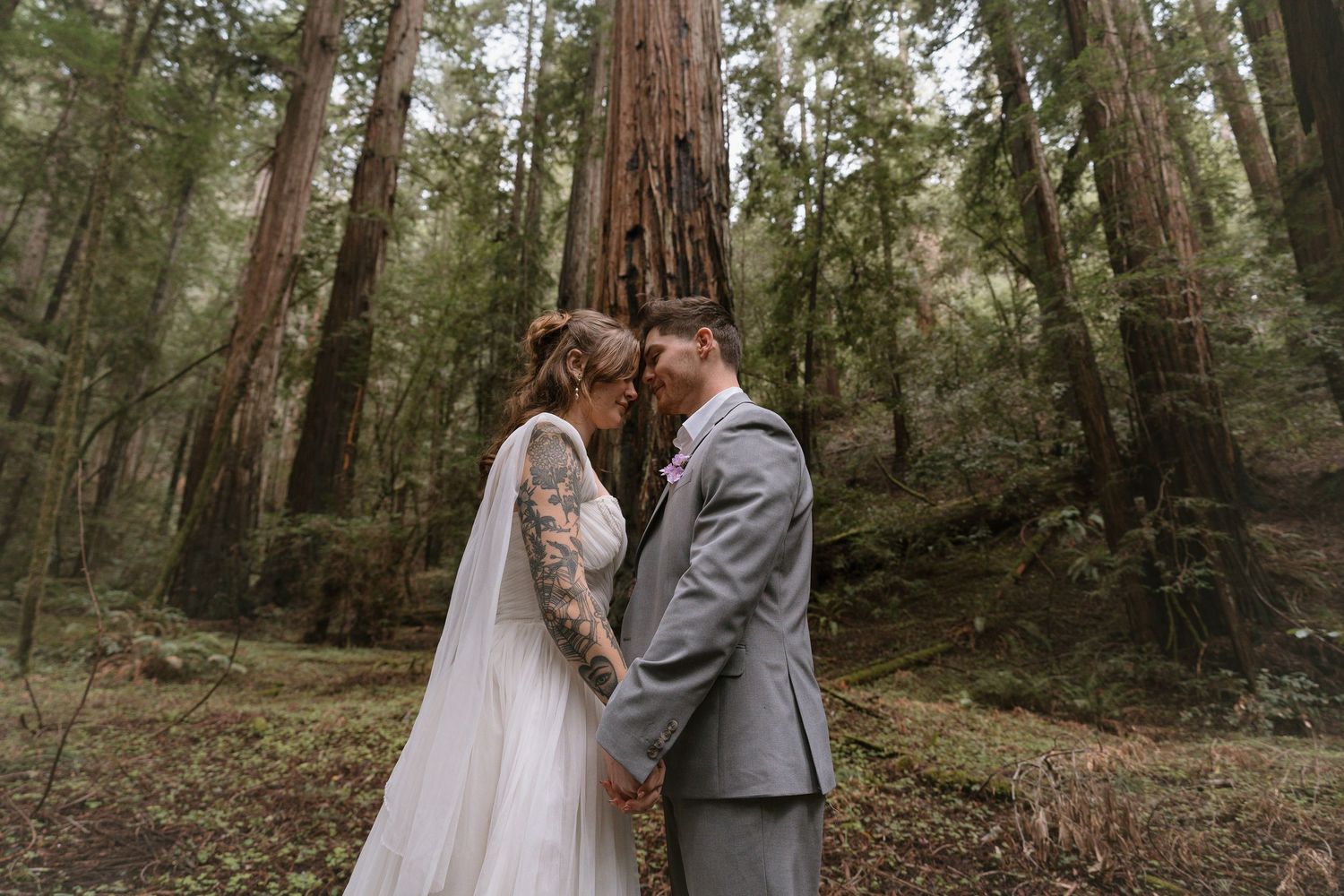 A couple in wedding attire walks through a serene redwood forest during their intimate outdoor ceremony.