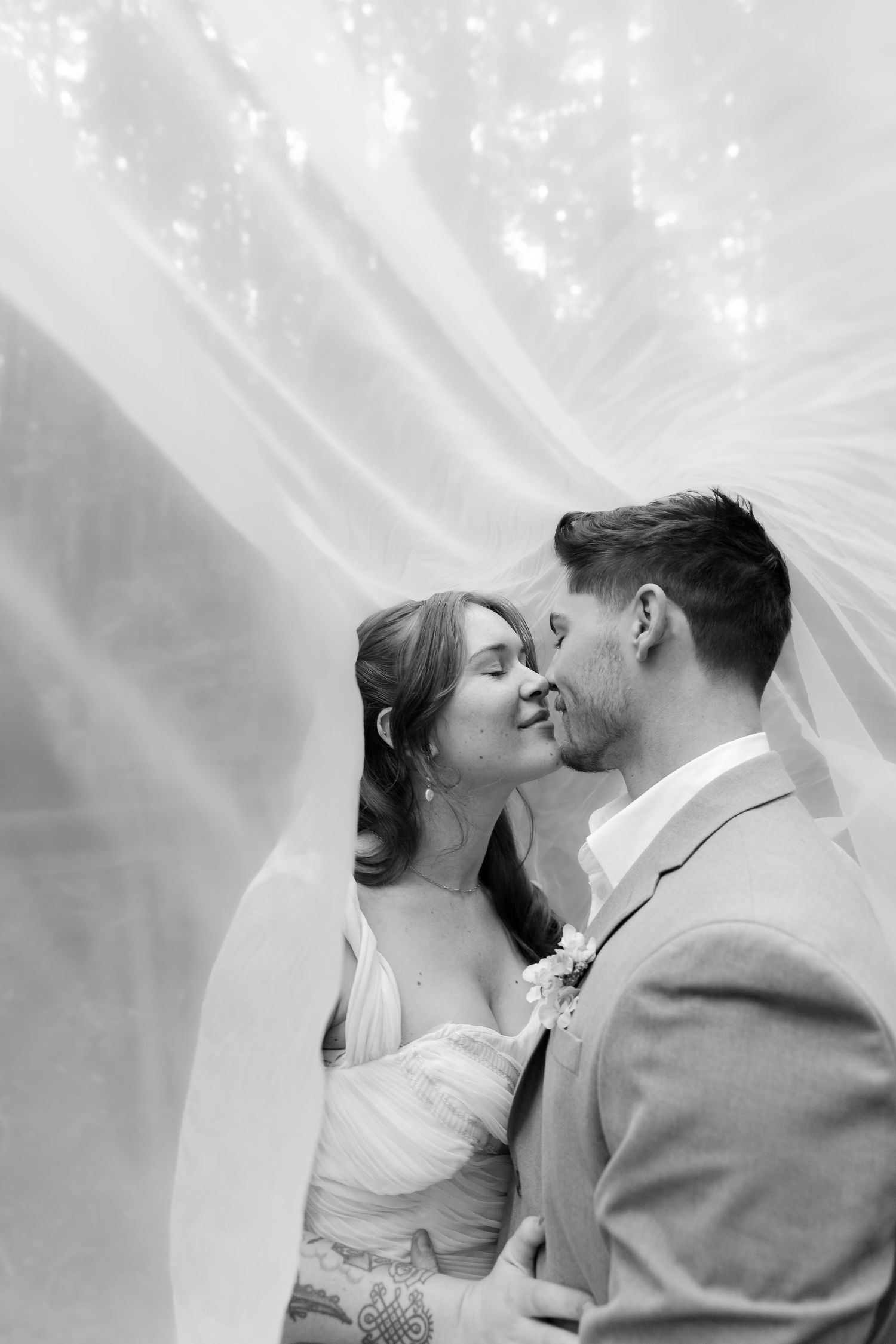 Black and white wedding photo of bride and groom sharing a tender moment under a flowing veil in a forest setting.