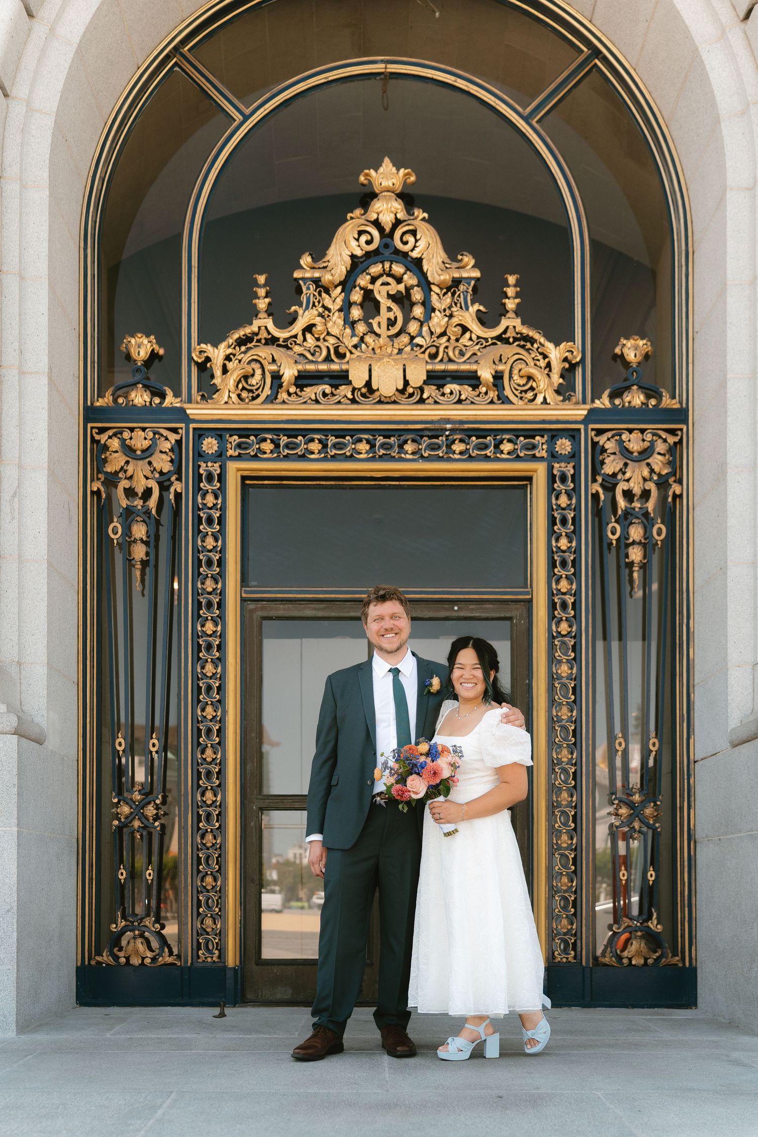 Couple in wedding attire poses before ornate gold San Francisco City Hall doors, bride holding colorful bouquet.