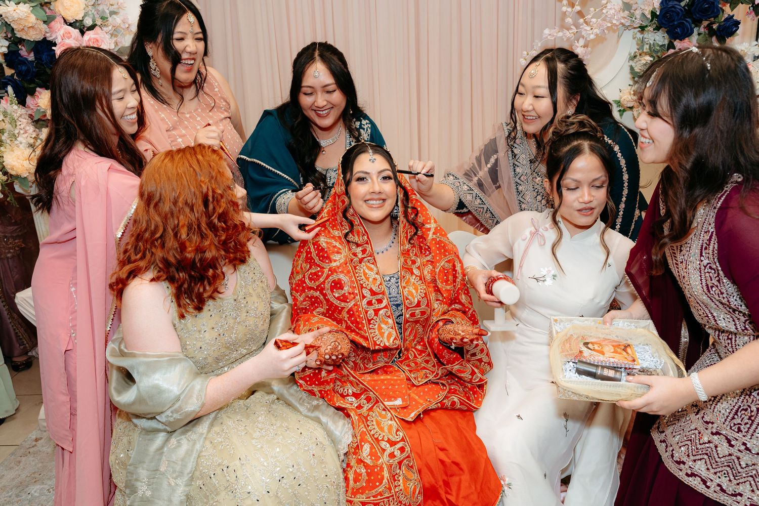 Bride in orange lehenga surrounded by bridesmaids during South Asian wedding mehndi ceremony.