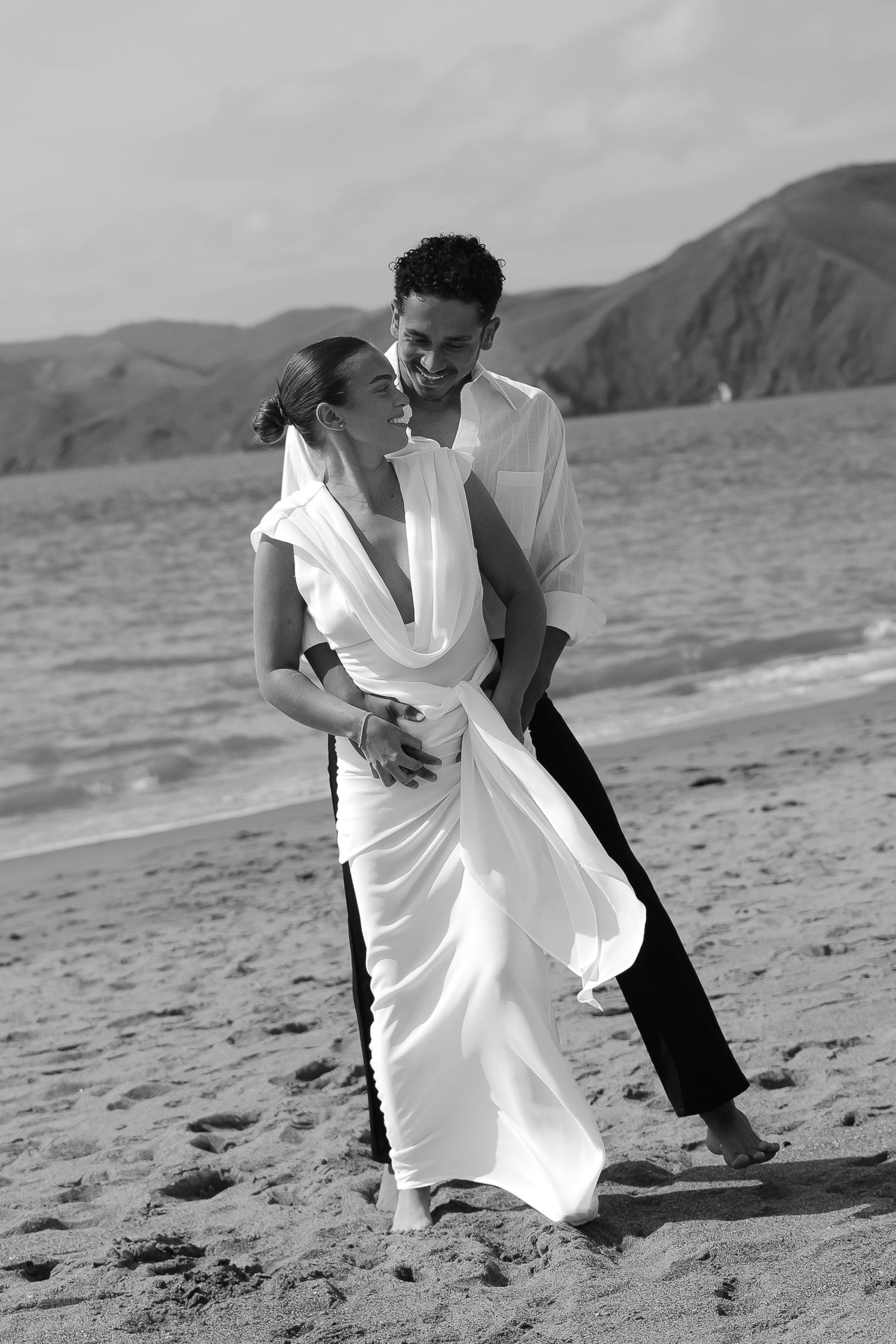 Couple in wedding attire embracing on a beach, woman in white gown and man in white shirt, black and white photo.