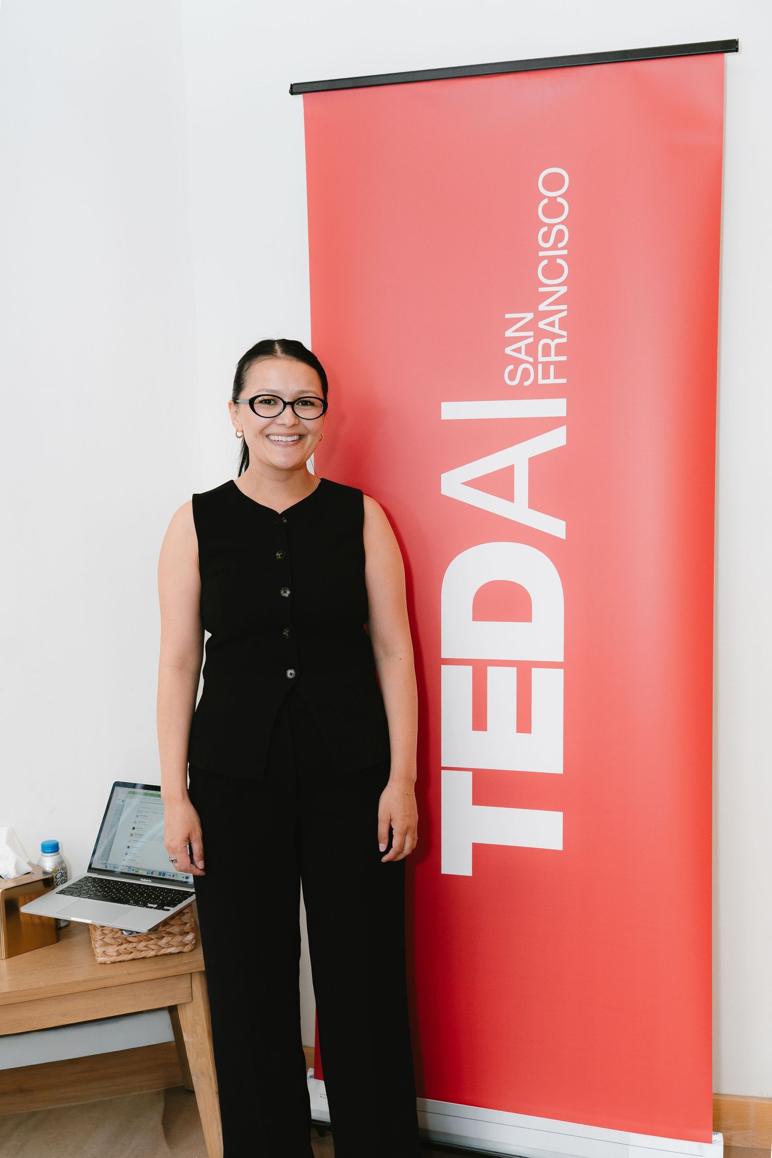 Woman in black outfit smiling beside a TEDai San Francisco banner at an indoor event.