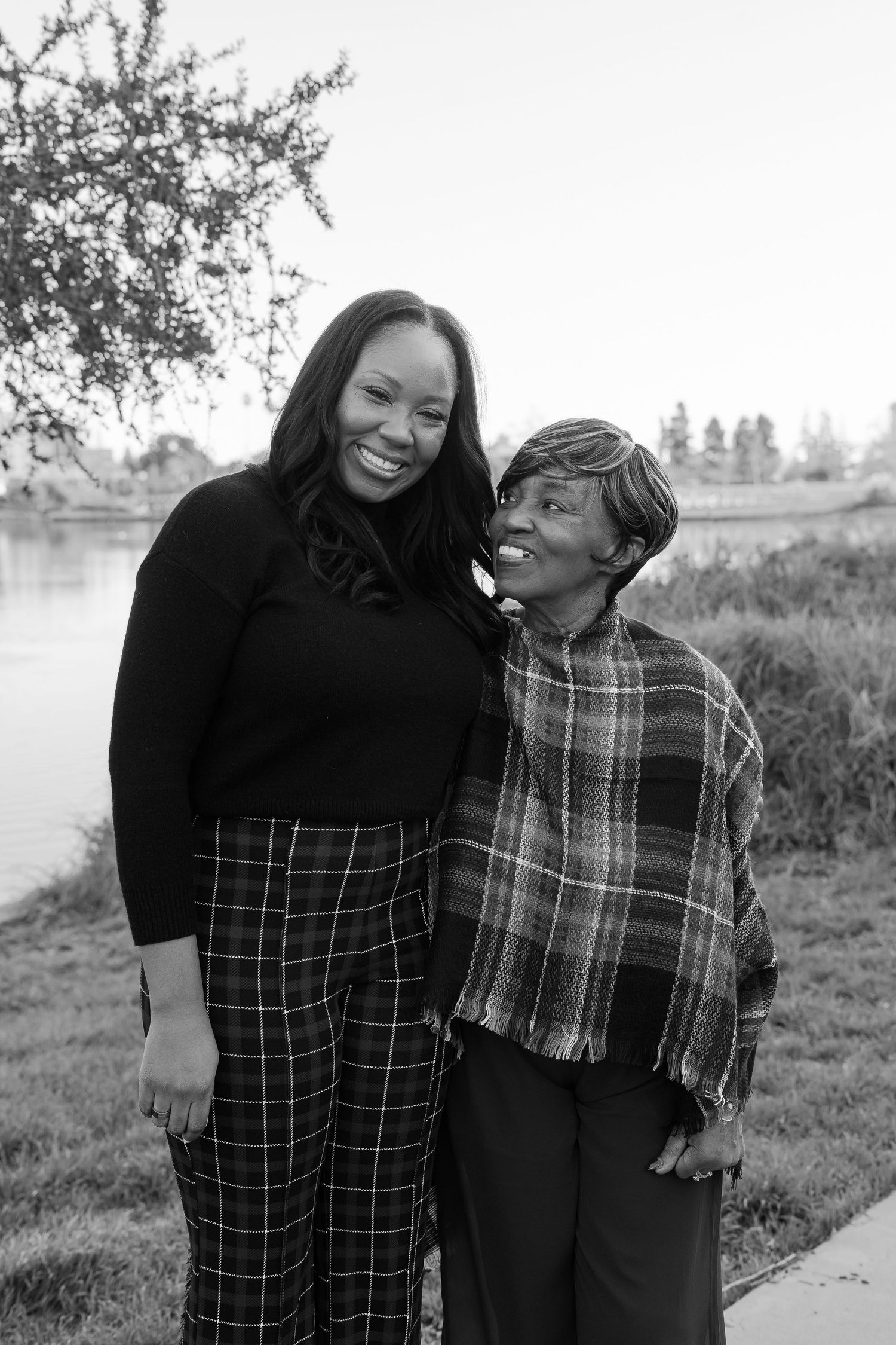 Two women smiling and embracing outdoors near a lake in a black and white portrait photo.