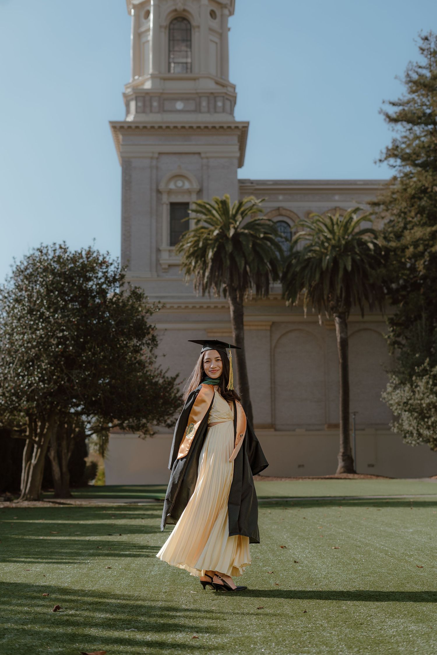 Young woman in graduation cap and gown poses on green lawn before a historic building with palm trees.