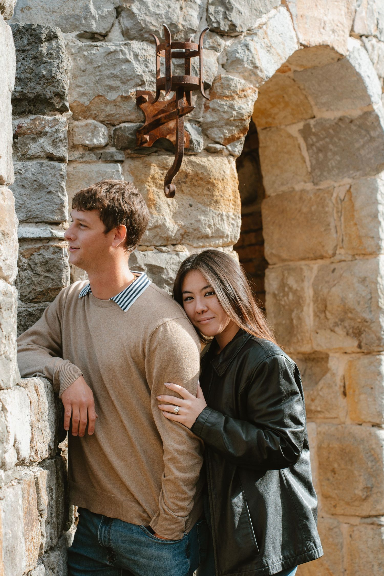 Couple in casual attire embracing against a rustic stone wall background.