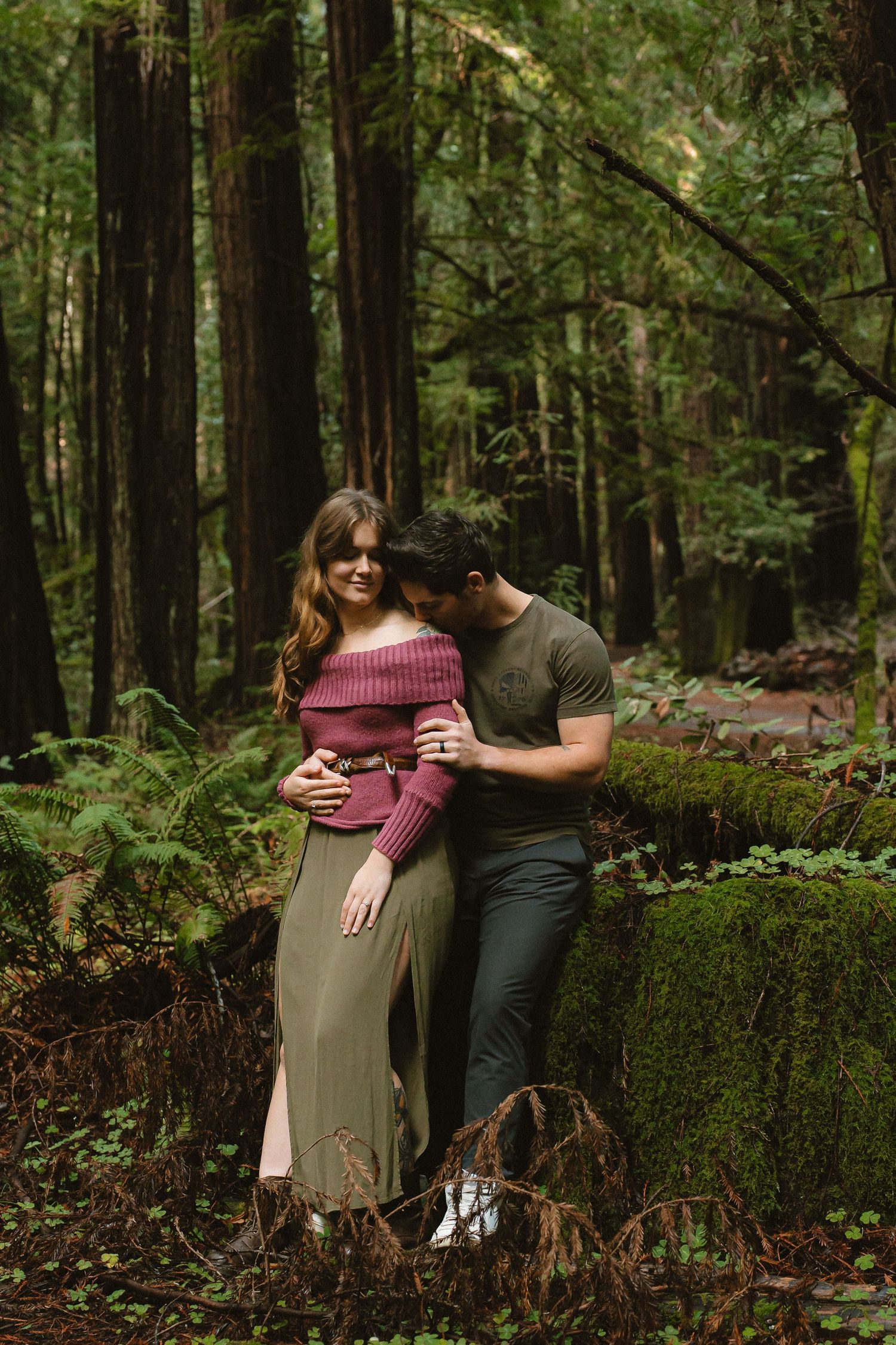 Couple embracing in a lush redwood forest, woman in pink off-shoulder top and olive skirt, man in green shirt.