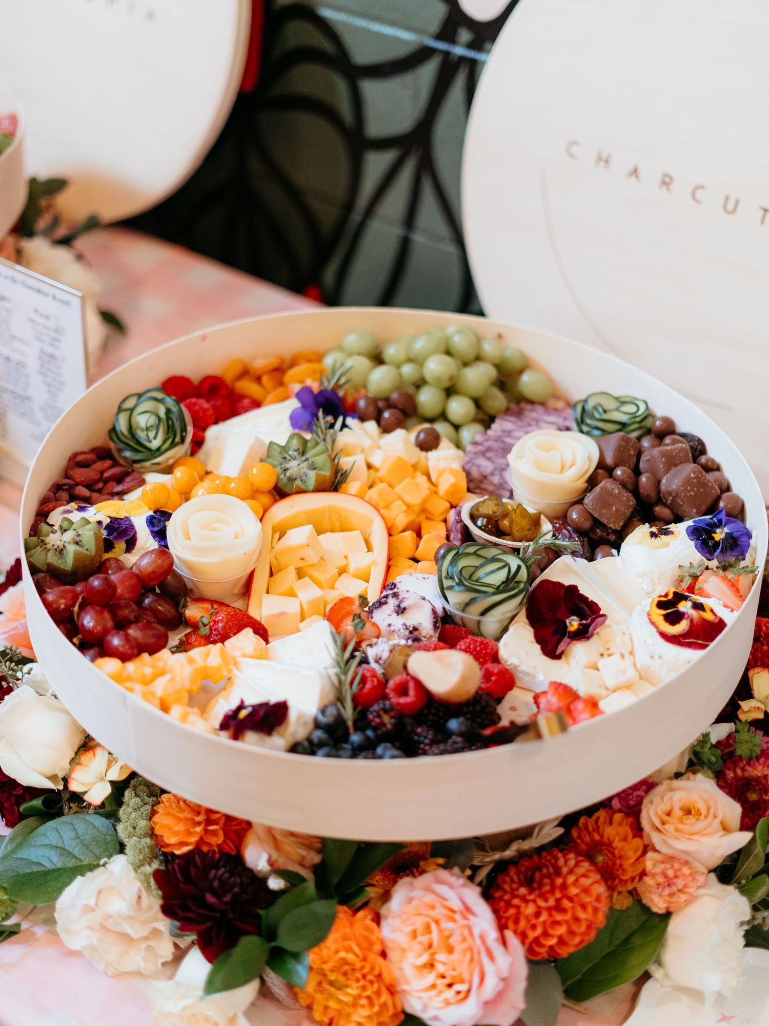 Elegant round charcuterie box filled with fruits, cheeses, chocolates, and flowers for a special event.