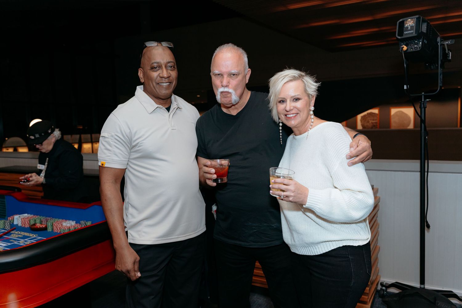 Three friends smile and pose together holding drinks at a casual indoor bar or bowling alley event.