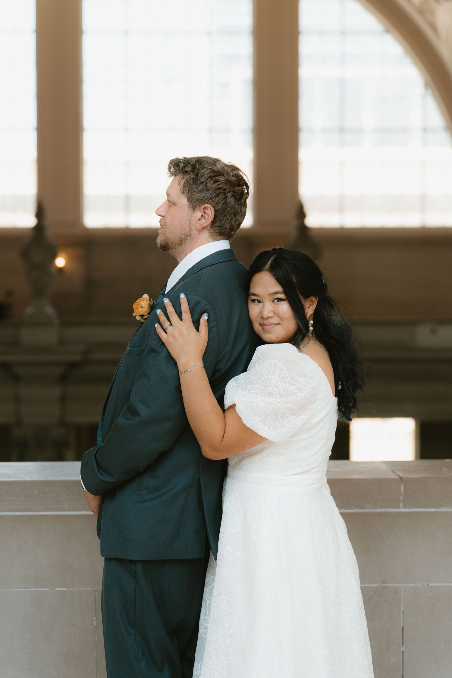 Asian bride embraces groom from behind near a sunlit window at San Francisco City Hall.