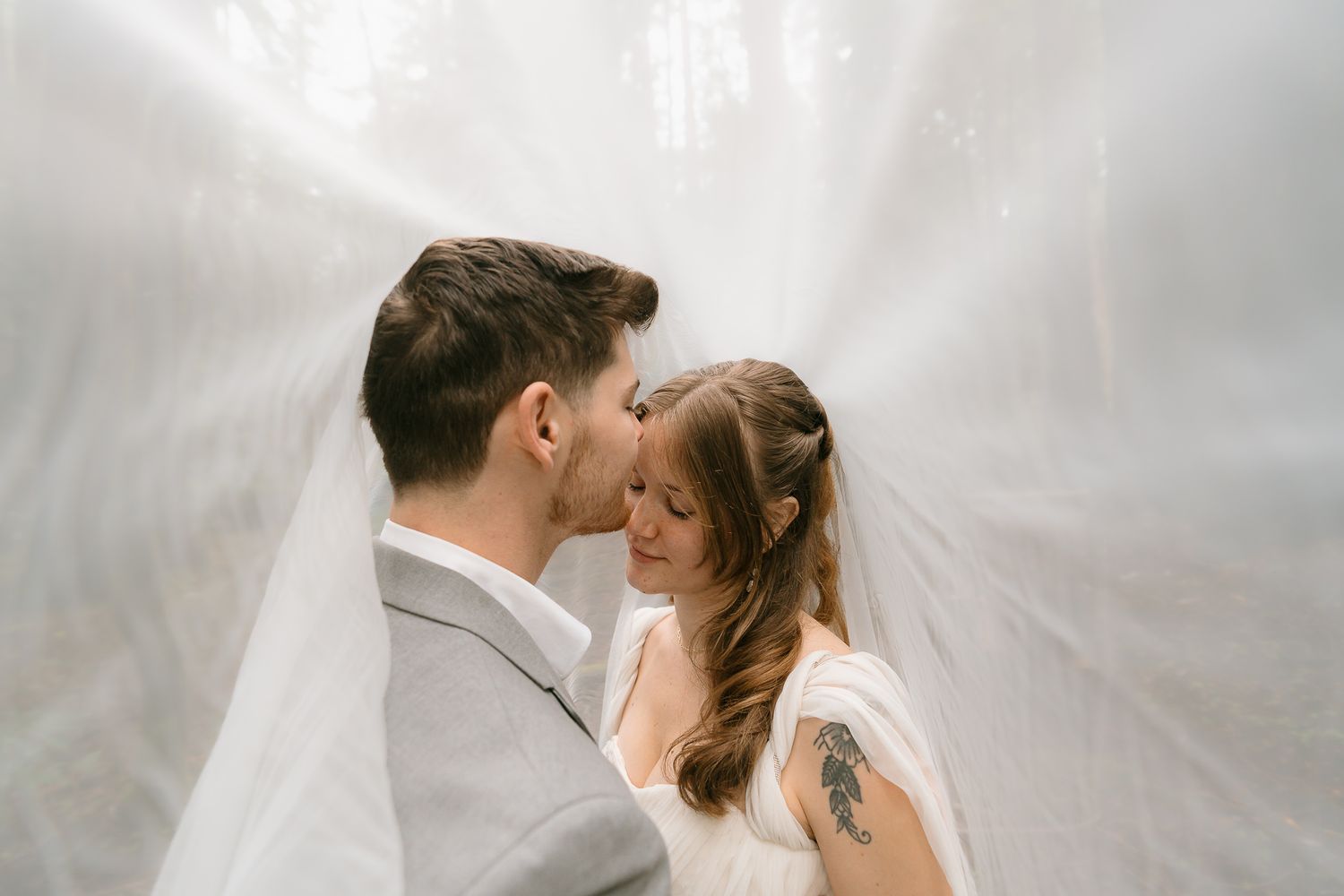 Bride and groom share romantic moment under flowing white veil in misty forest wedding photo.