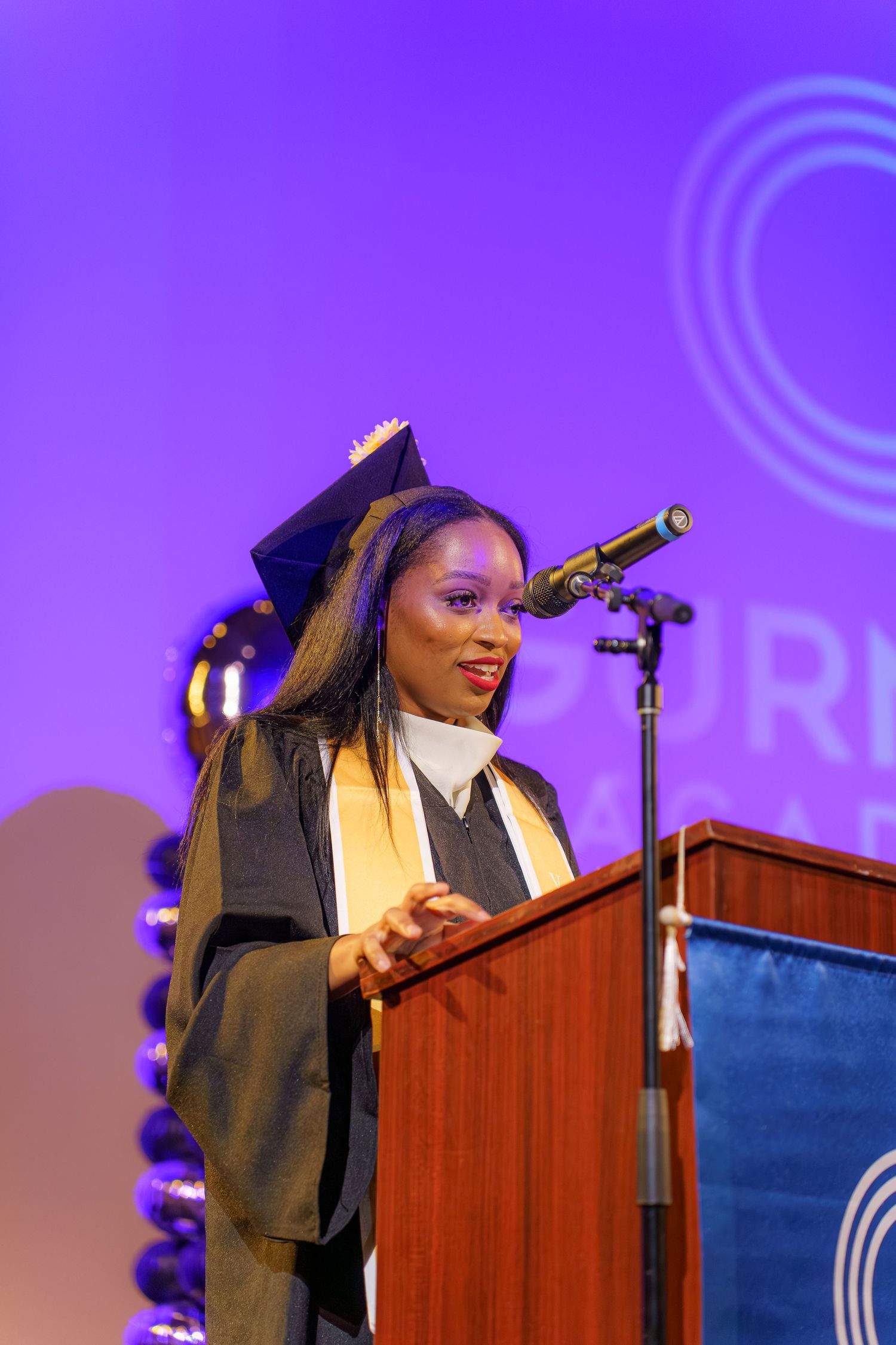 Graduate in cap and gown delivers commencement speech at podium during graduation ceremony with purple backdrop.