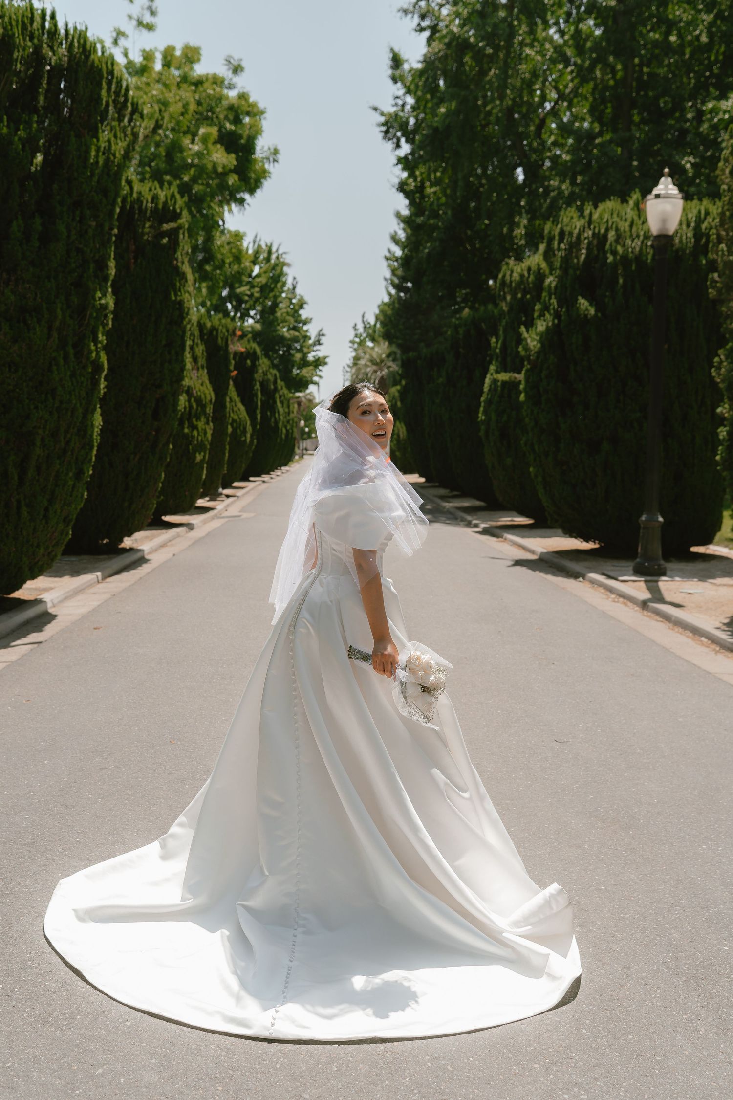 Elegant bride in long white gown and veil poses on tree-lined garden pathway.
