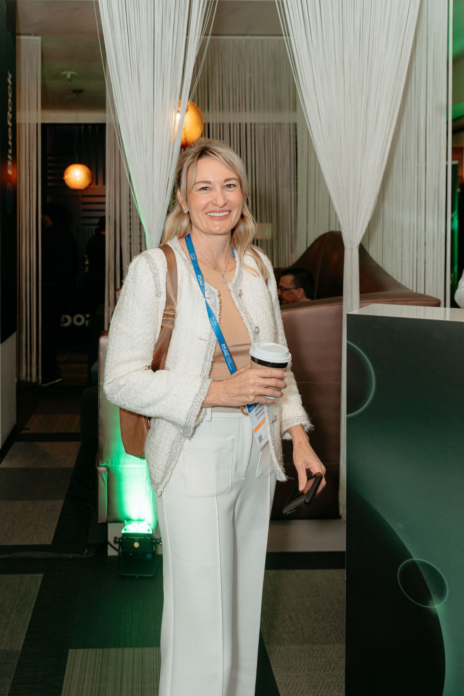 Smiling blonde woman in white blazer and pants holding coffee cup at an indoor event venue.