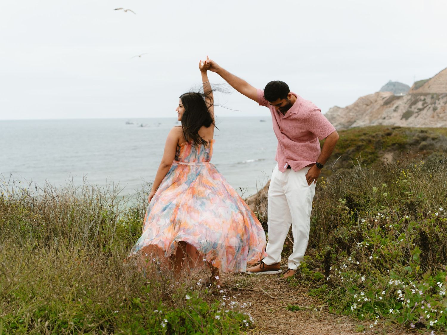 A couple dances on a coastal cliff, woman in colorful floral dress, ocean views behind them.