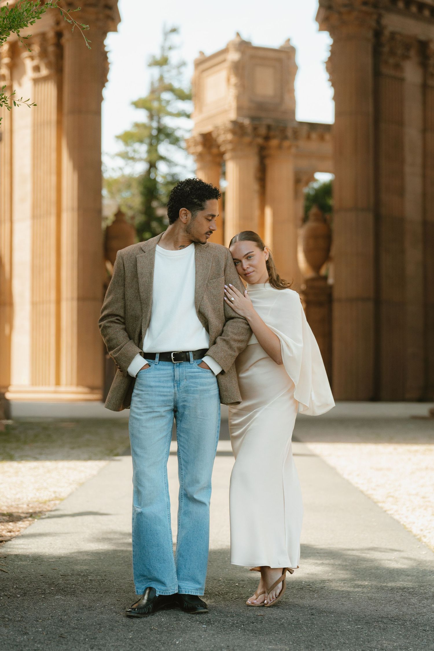 Couple posing at Palace of Fine Arts, woman in white dress leaning on man in blazer and jeans, romantic outdoor portrait.