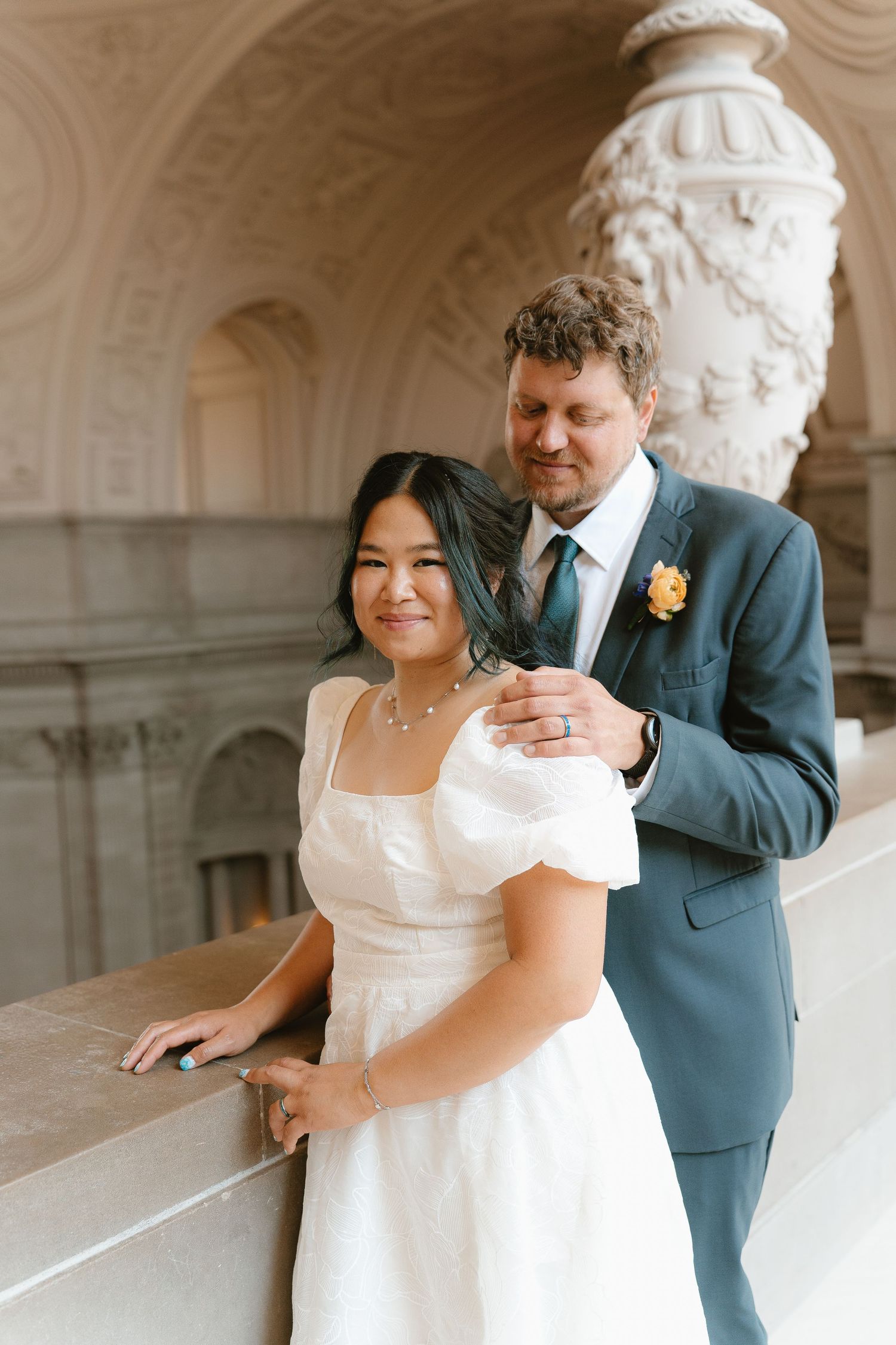 Groom in teal suit embraces bride in white dress against ornate stone architecture at San Francisco City Hall.