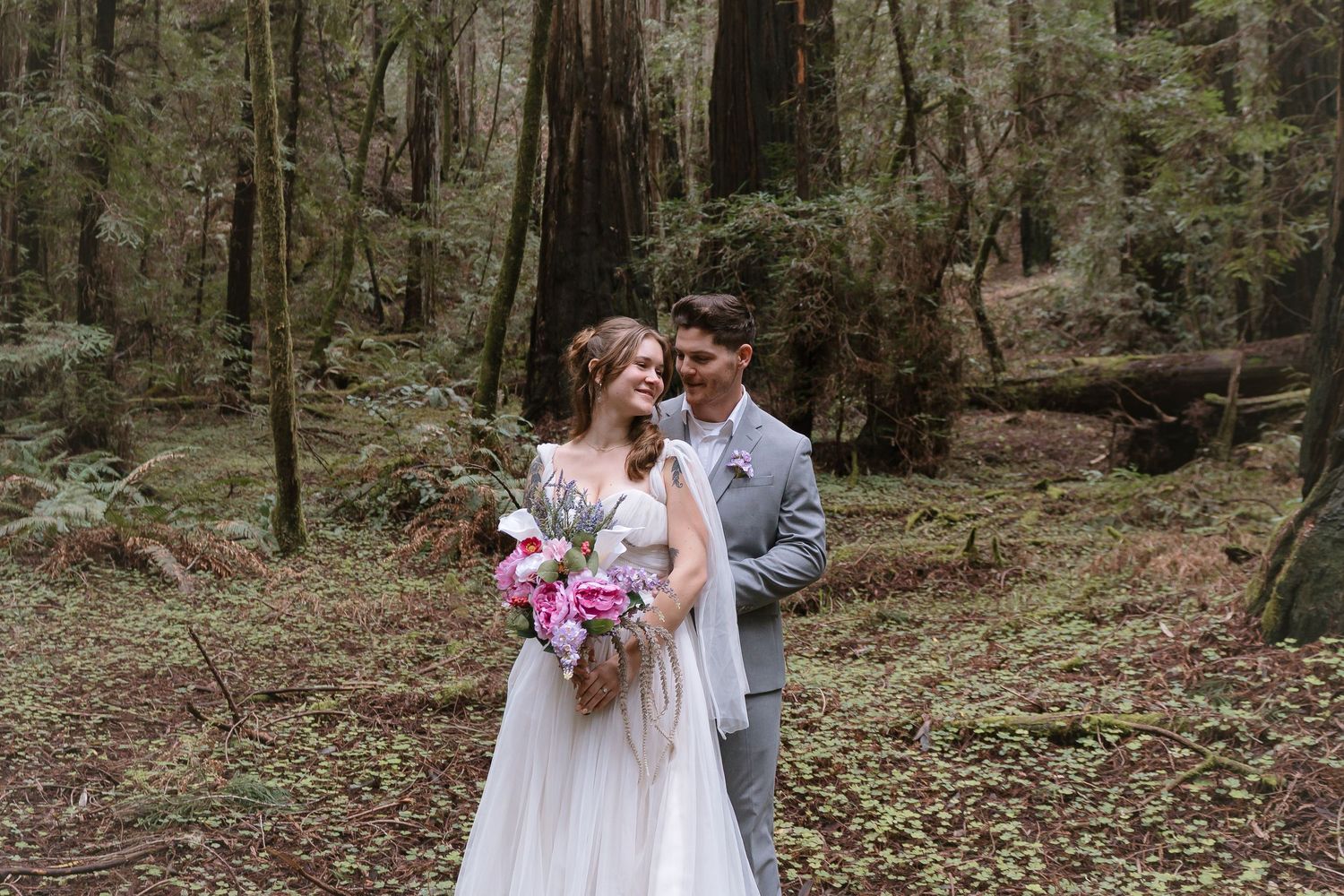 Bride and groom embracing in a lush redwood forest, bride holding pink floral bouquet in a flowing white gown.