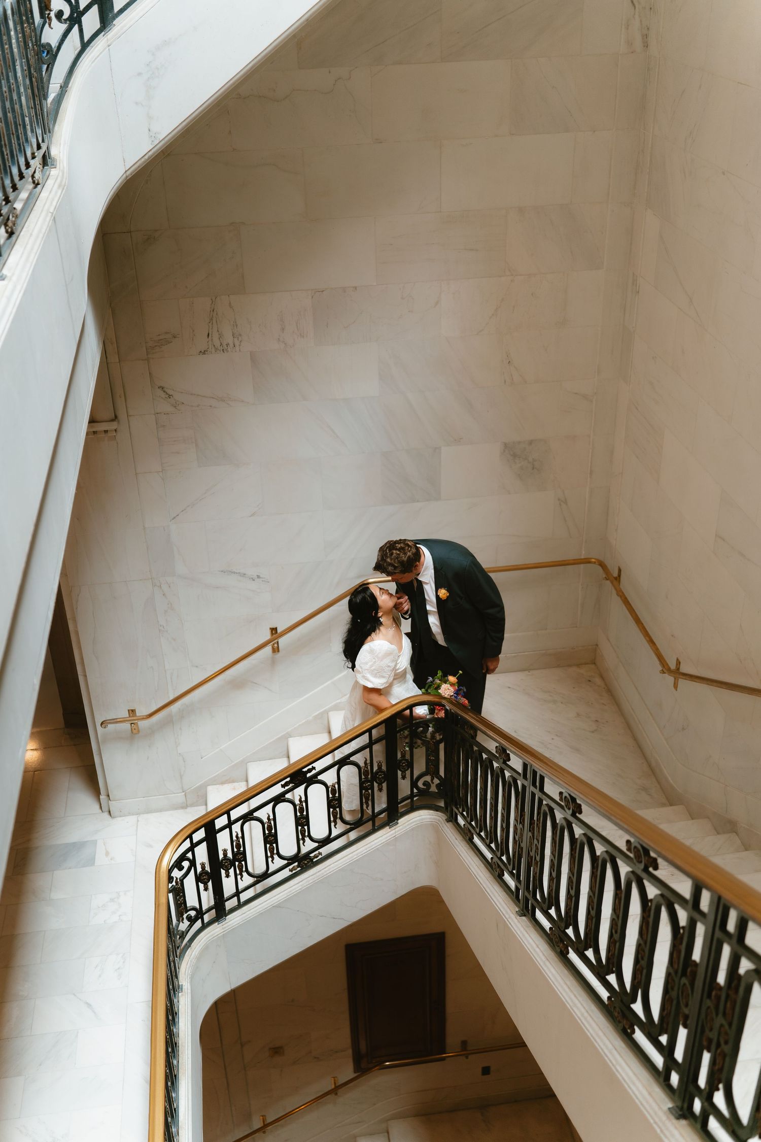 Newlyweds share a romantic kiss on a marble staircase with ornate iron railings.