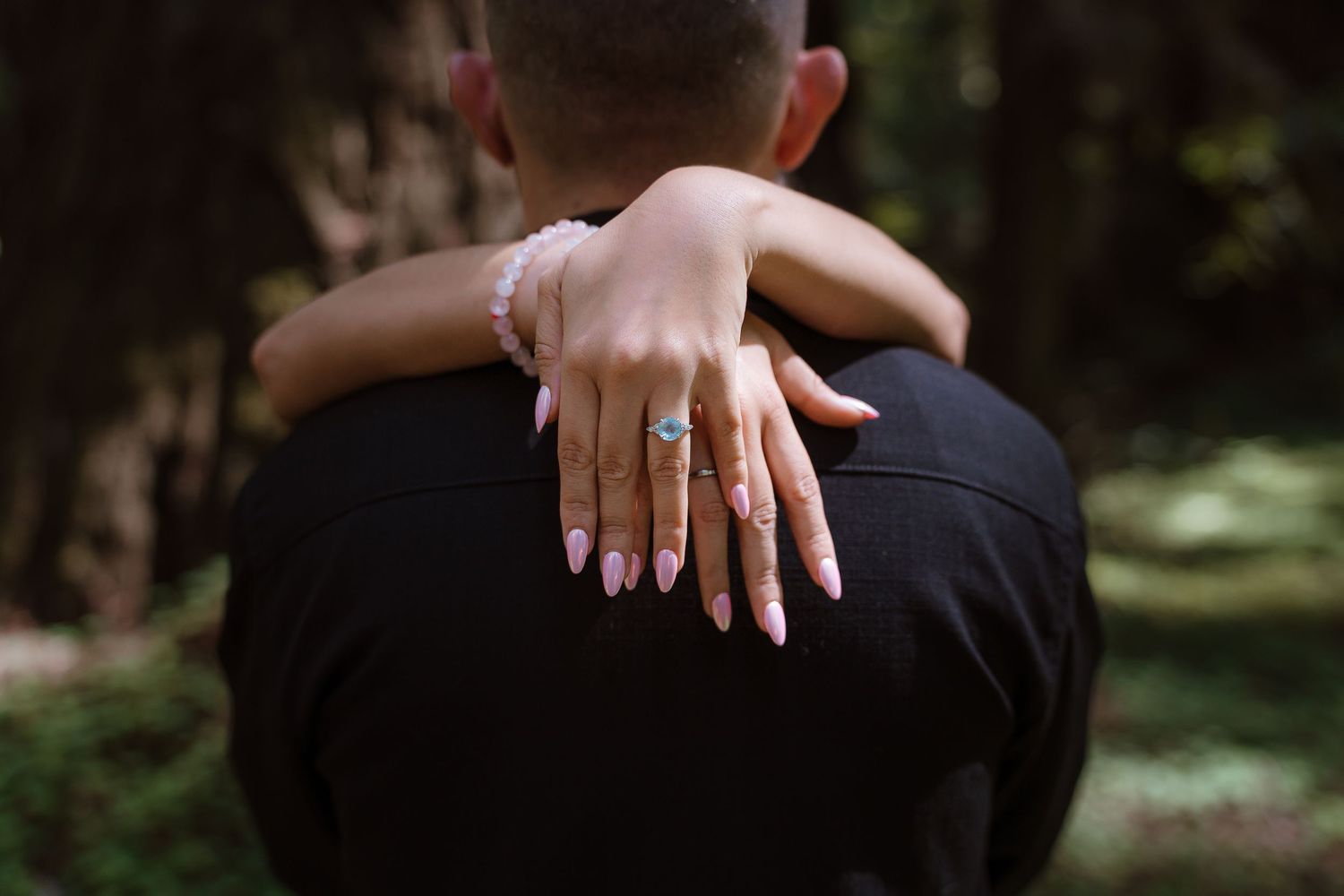 Woman with pink nails and blue gemstone ring embraces man in black shirt outdoors in forest setting.