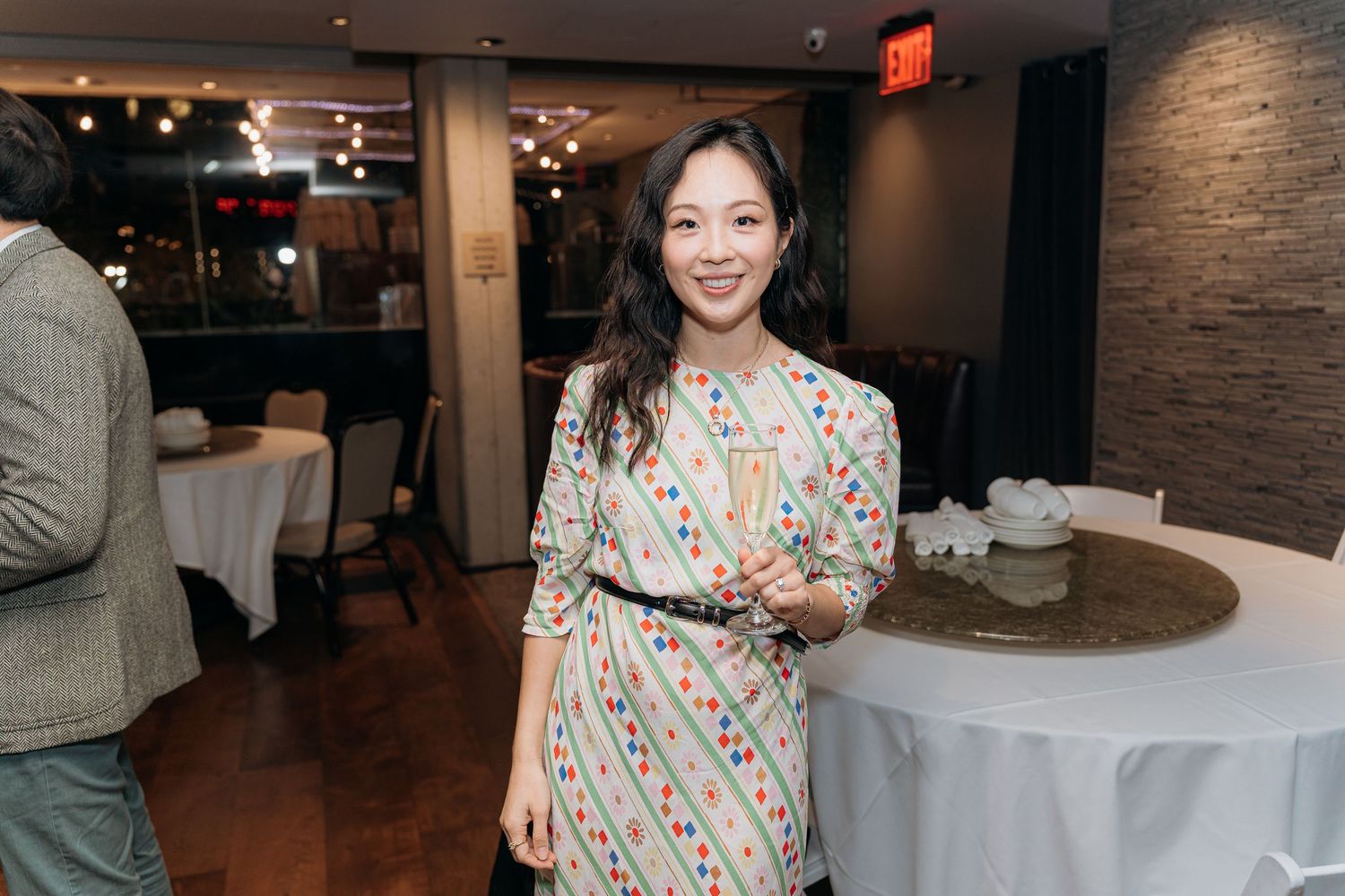 Major Milestone birthday where Woman in colorful patterned dress smiling at indoor restaurant event with white tablecloths.