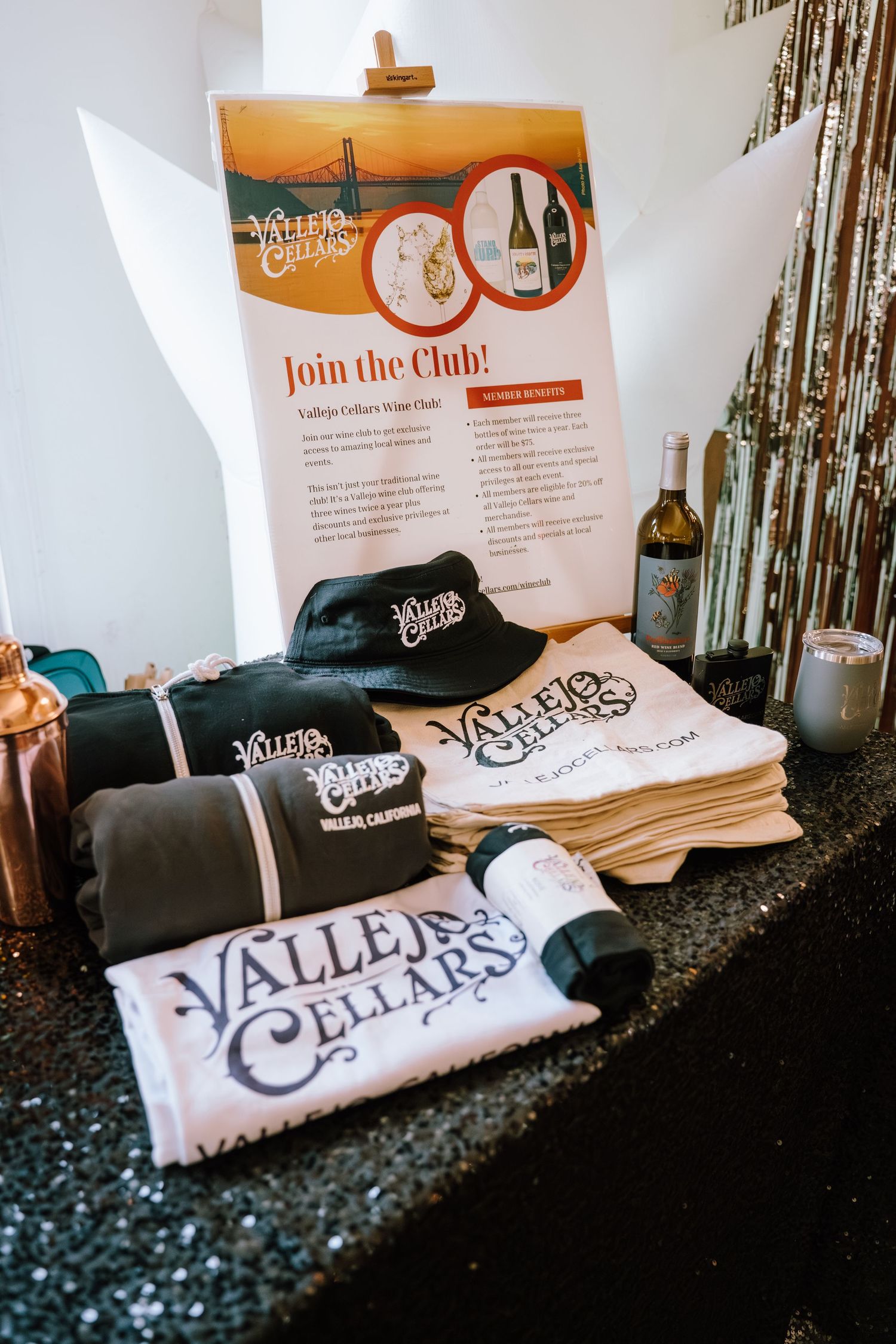 Vallejo Cellars wine club merchandise display featuring branded t-shirts, hats, and wine bottles on a glittery black table.