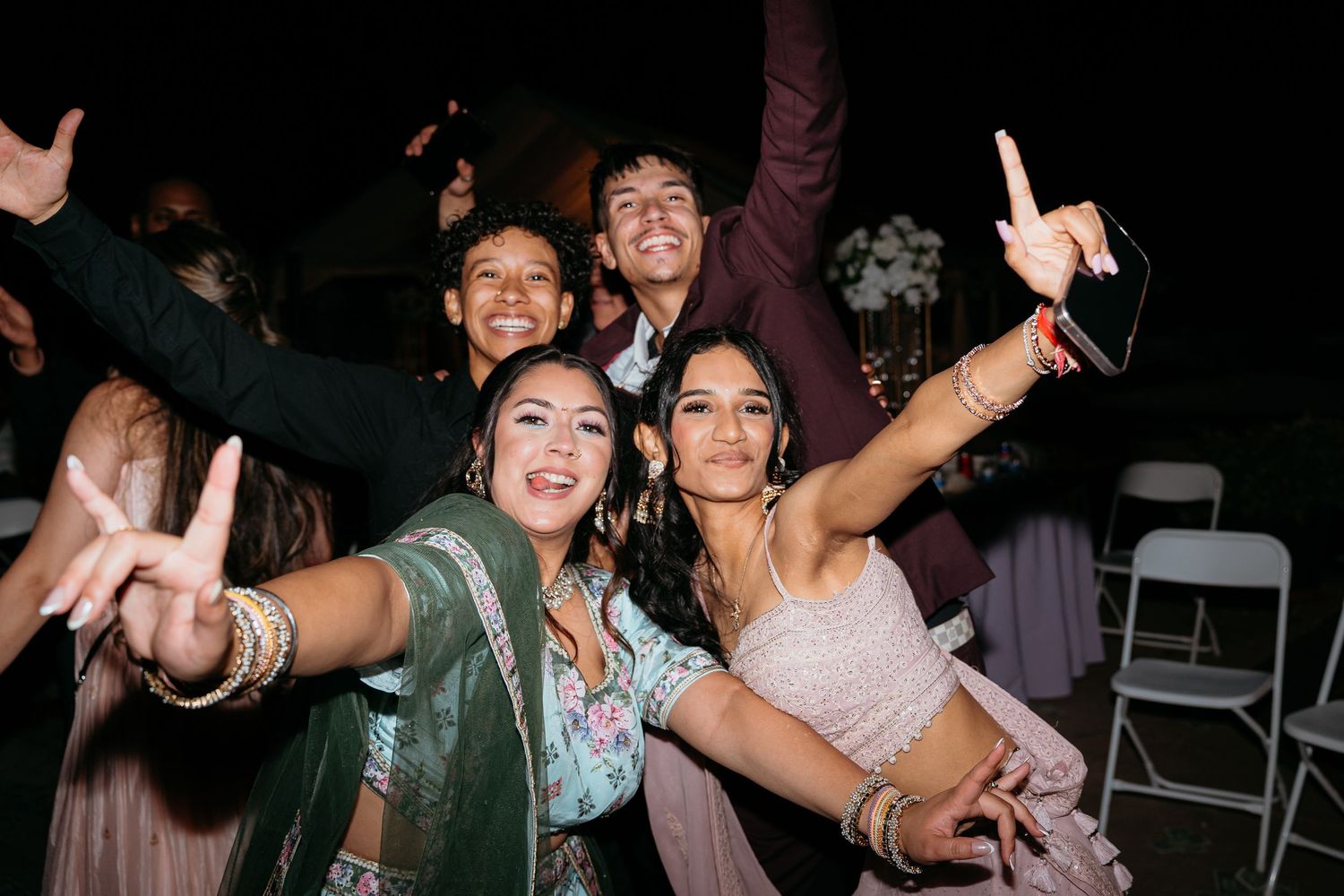 Group of friends in traditional Indian attire celebrating at a wedding reception, making peace signs and dancing outdoors at night.