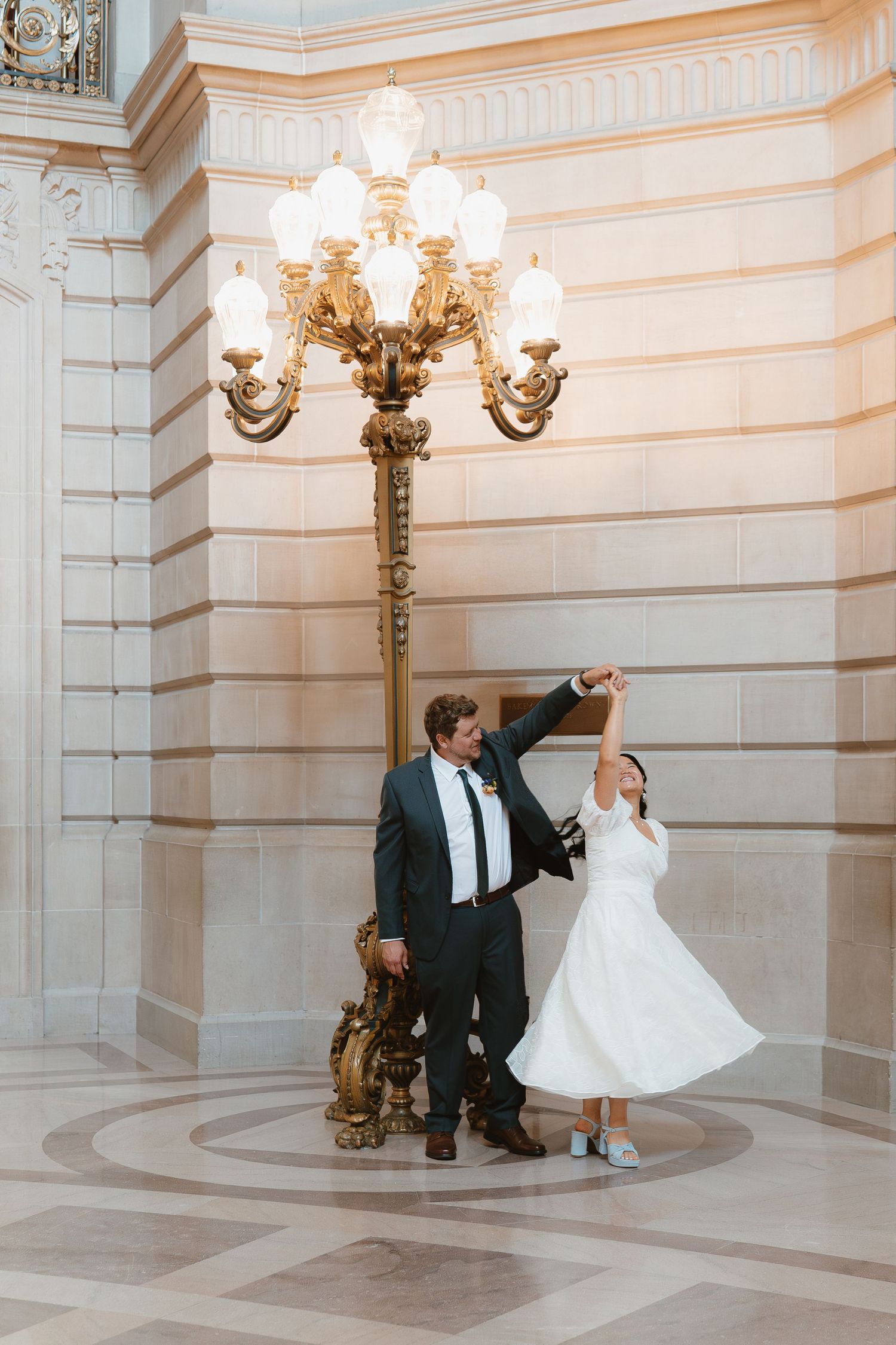 Bride and groom dancing under ornate gold lamp post in San Francisco City Hall rotunda.