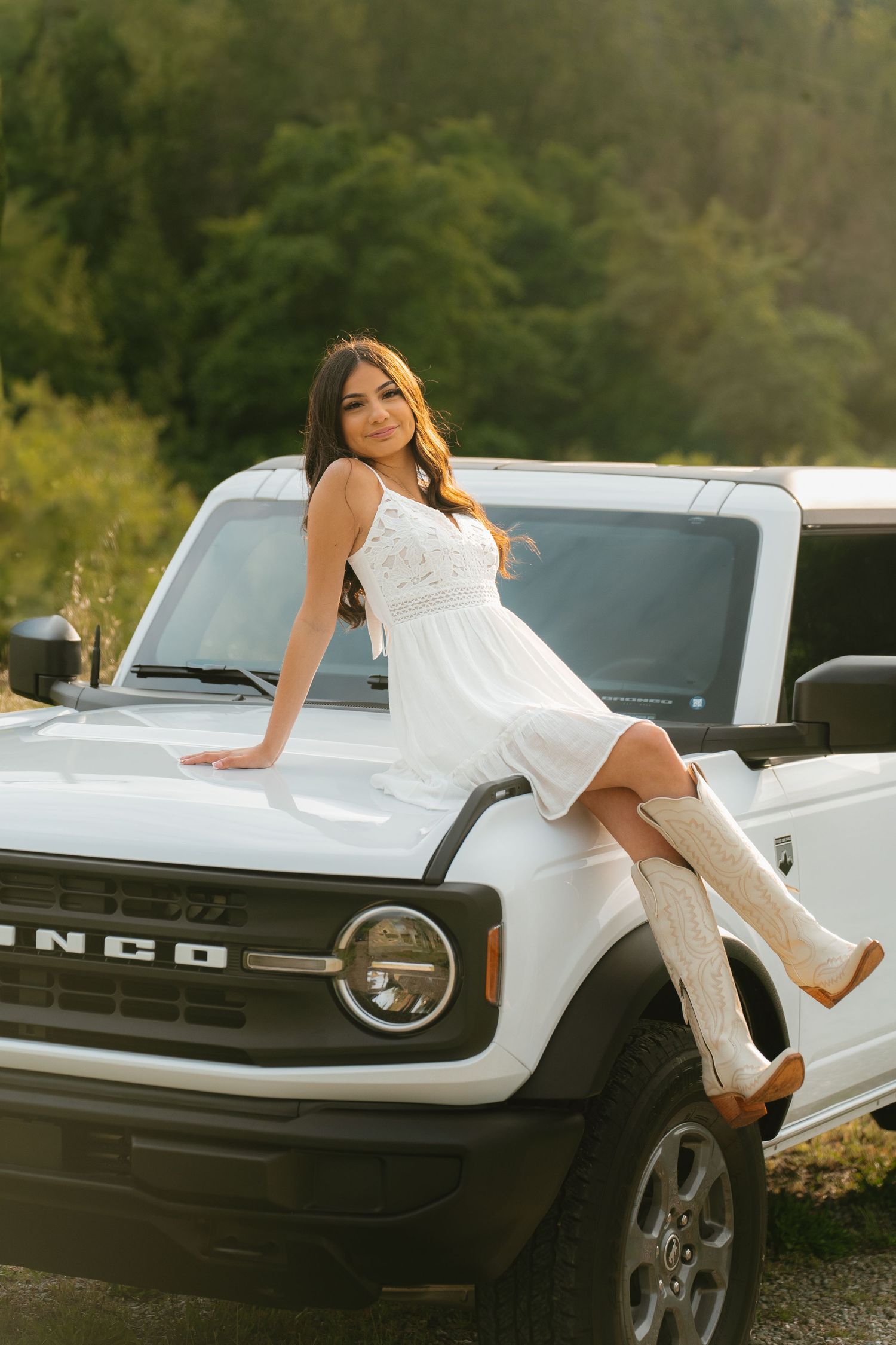 Young woman in white dress and cowboy boots posing on hood of white Ford Bronco outdoors.
